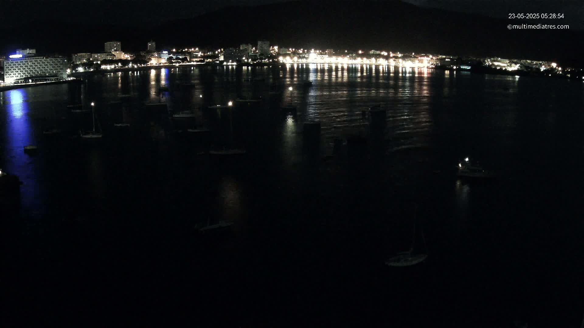 A dark nighttime view of a calm body of water with numerous boats moored, reflecting the lights of a city on the far shore.