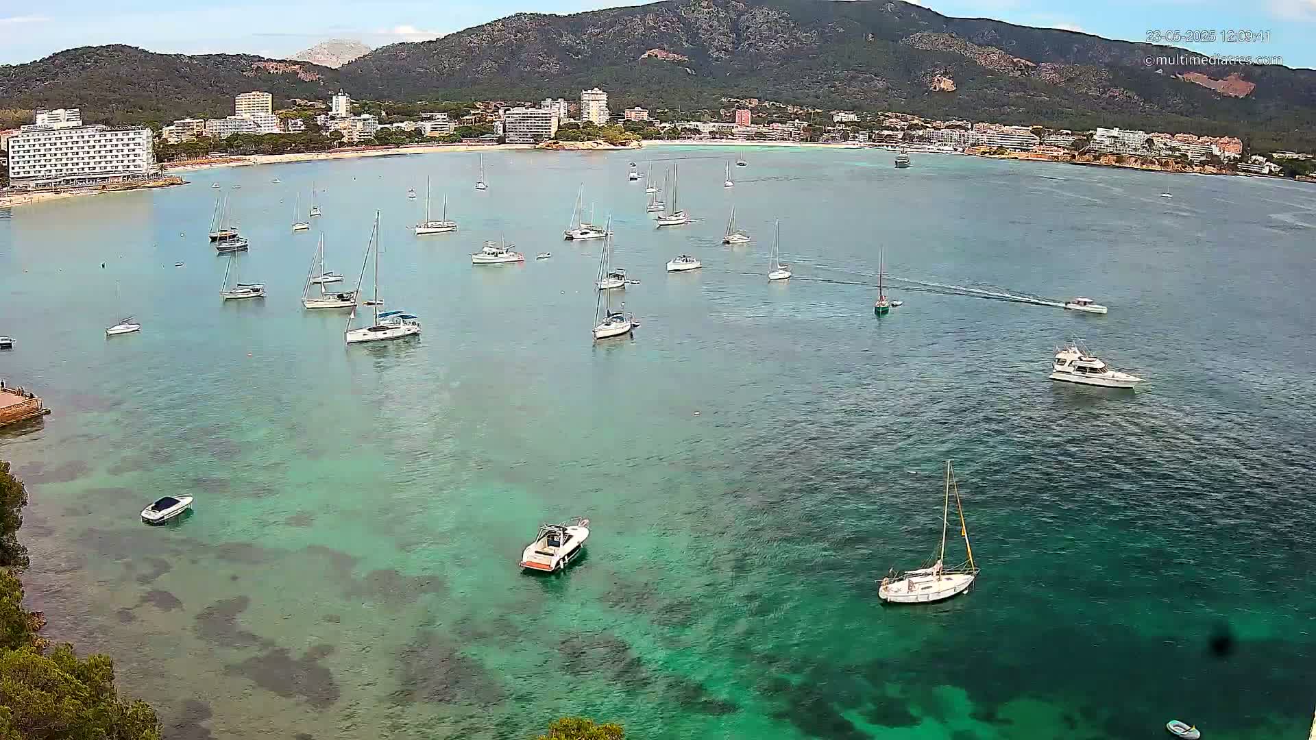 A calm, clear turquoise bay with numerous sailboats and motorboats anchored near a beach and a mountainous coastline under a partly cloudy sky.