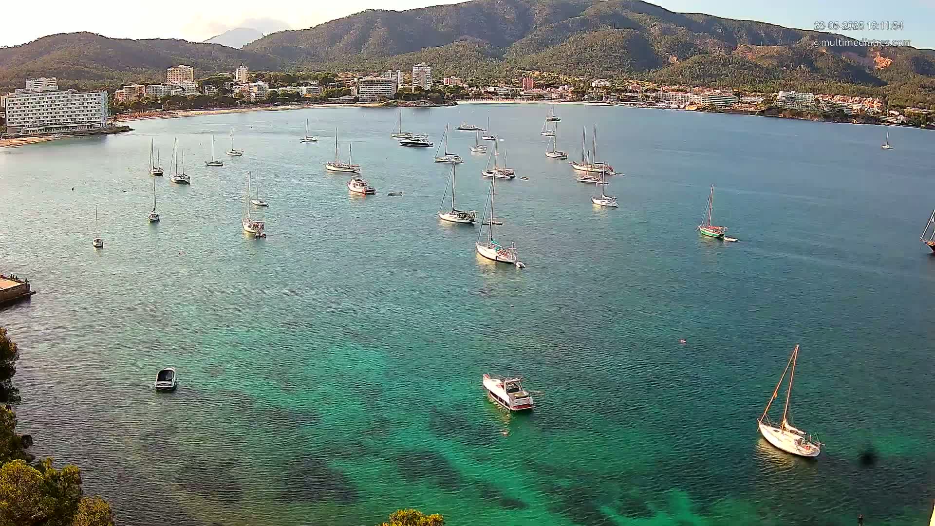 A calm, clear bay with numerous sailboats and motorboats is nestled against a shoreline of hotels and lush green mountains under a bright sky.