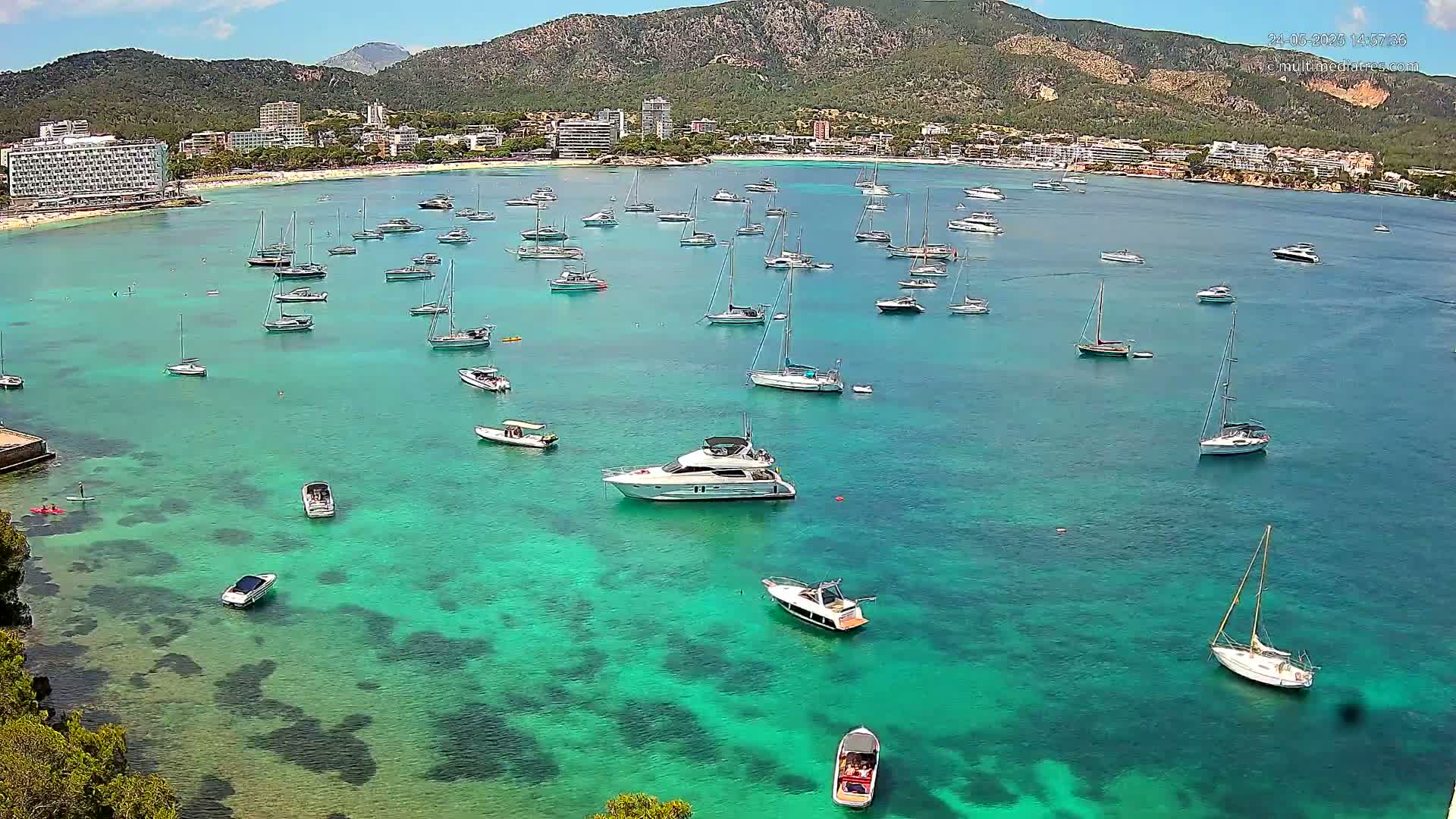 A calm, sunny day shows numerous sailboats and motorboats anchored in a clear, turquoise bay, with a resort town and mountains visible in the background.