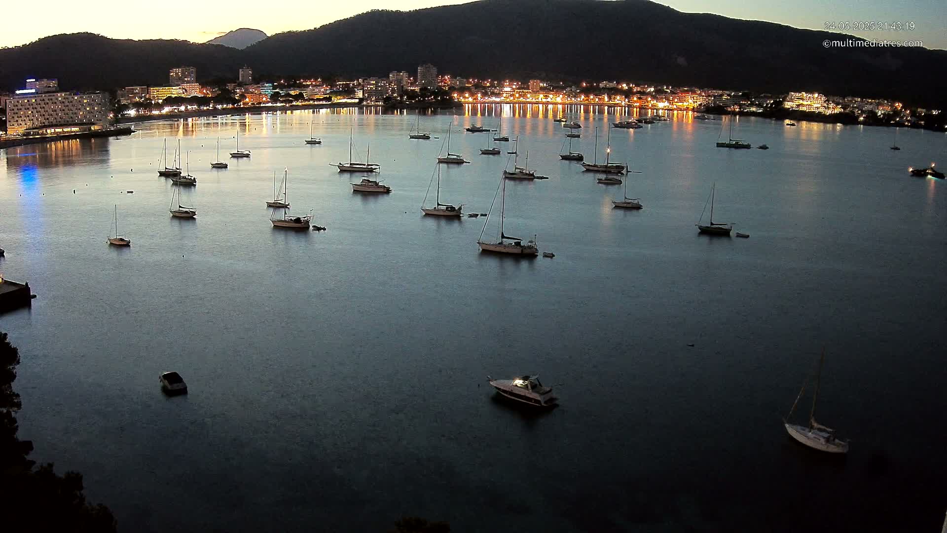 Numerous sailboats are moored in a calm, dark bay at twilight, with a brightly lit town visible on the far shore.