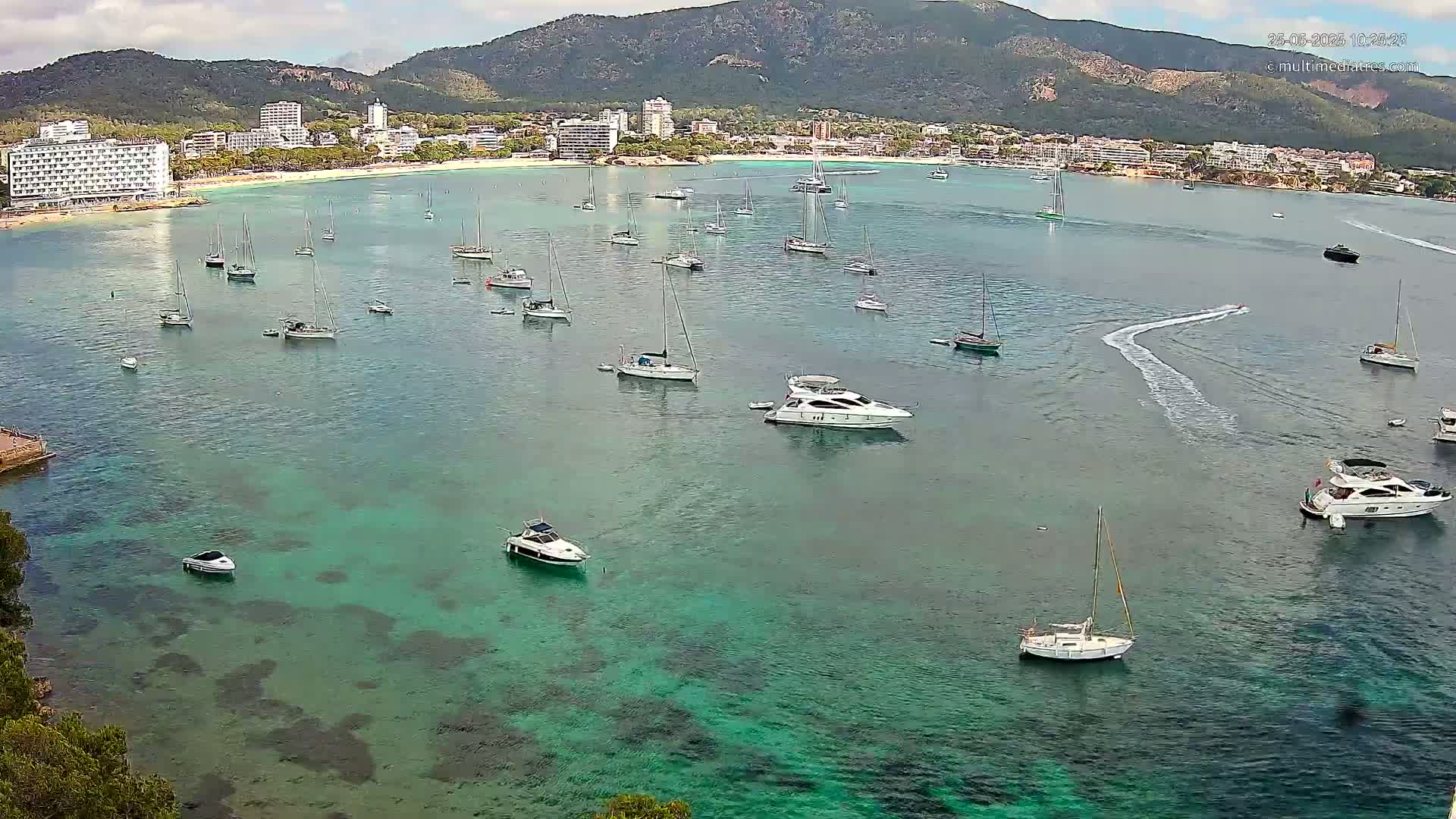 A calm, clear turquoise bay with numerous sailboats and motorboats anchored near a shoreline of buildings and a mountainous backdrop under a partly cloudy sky.