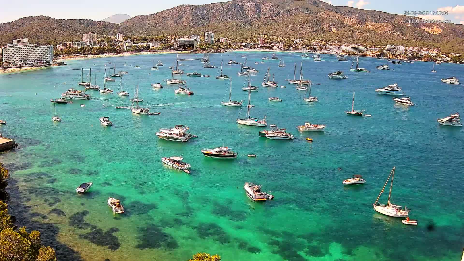 A sunny day shows numerous boats and yachts anchored in a clear, turquoise bay near a coastline with buildings and mountains in the background.
