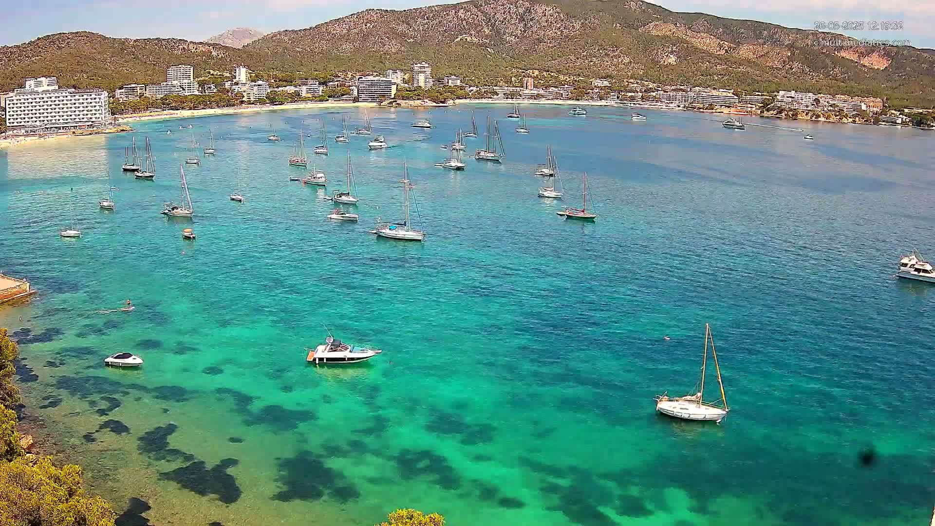 Numerous sailboats are anchored in a calm, clear turquoise bay near a beach lined with hotels and a mountainous backdrop under sunny skies.