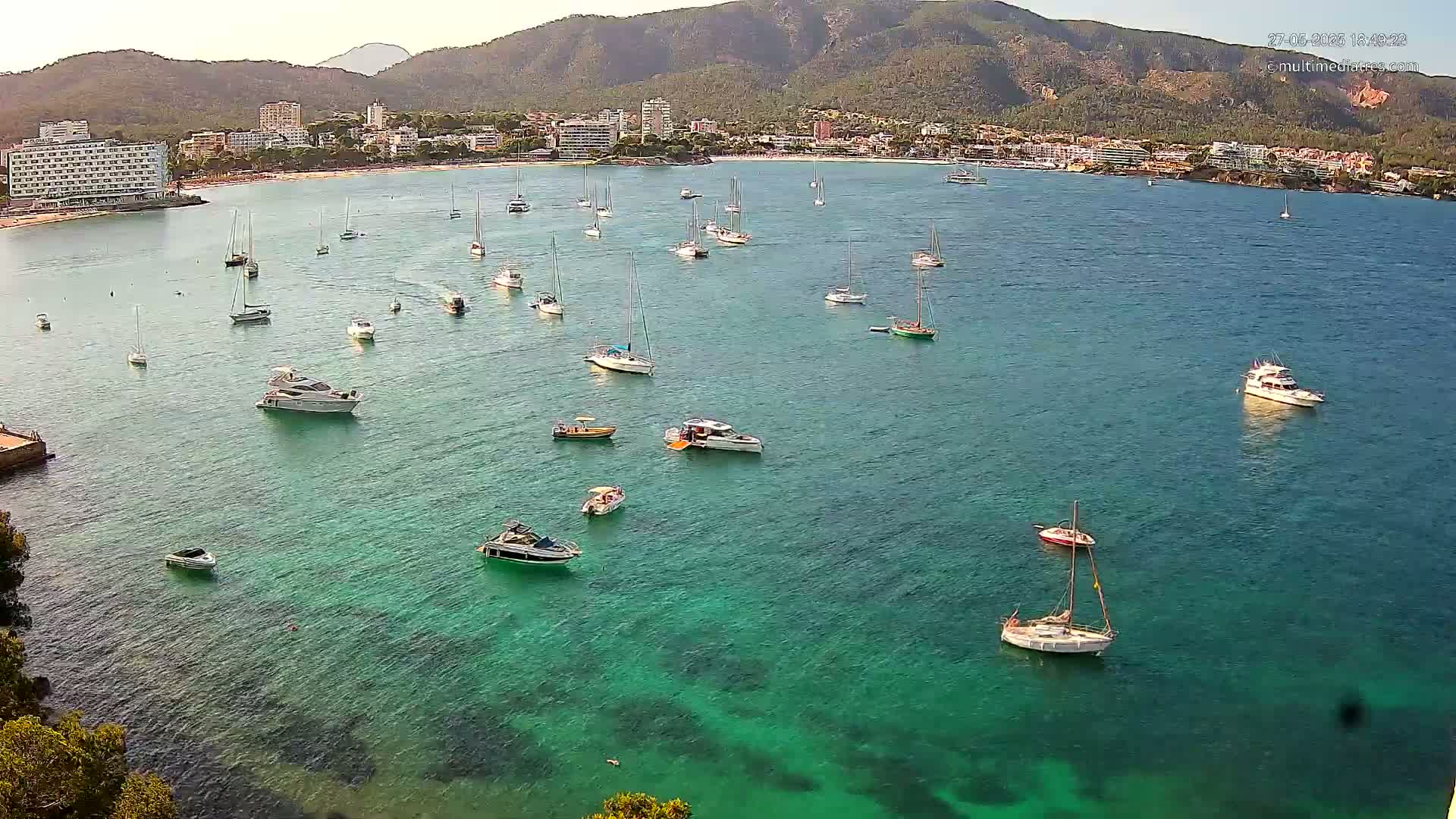Numerous boats and yachts are anchored in a calm, clear turquoise bay, with a mountainous coastline and buildings in the background under a sunny sky.