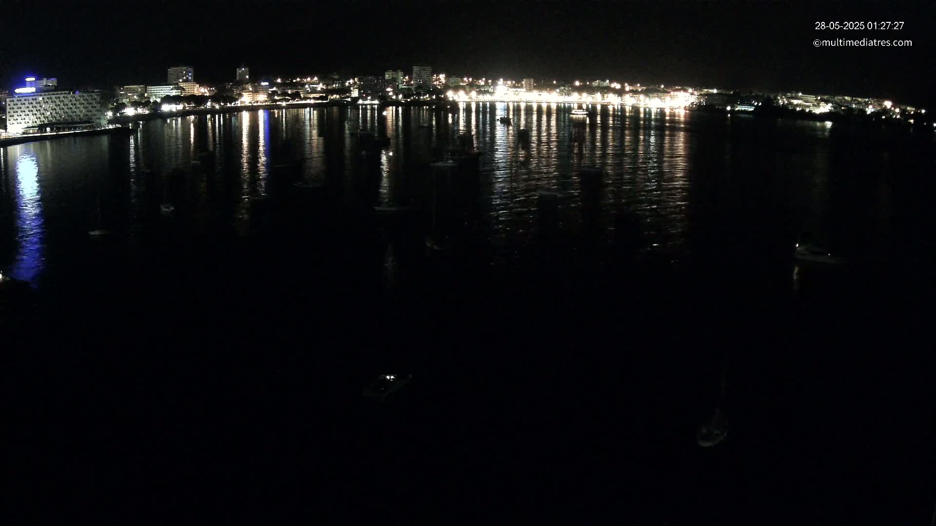 A nighttime view of a calm body of water reflecting the lights of a city skyline across its surface, with several small boats scattered throughout.