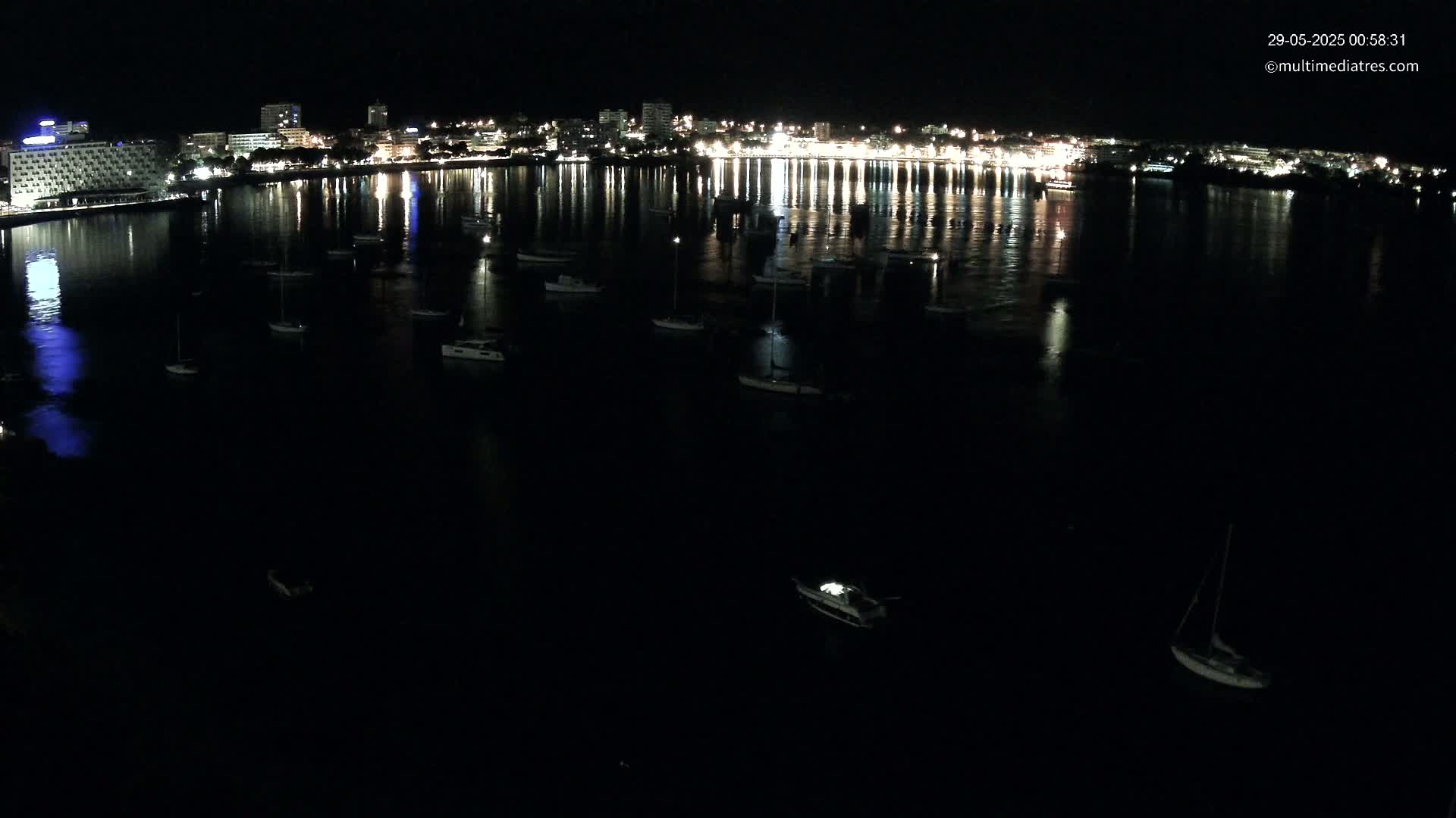 A nighttime view of a calm body of water with numerous boats moored, reflecting the lights of a city skyline in the background.
