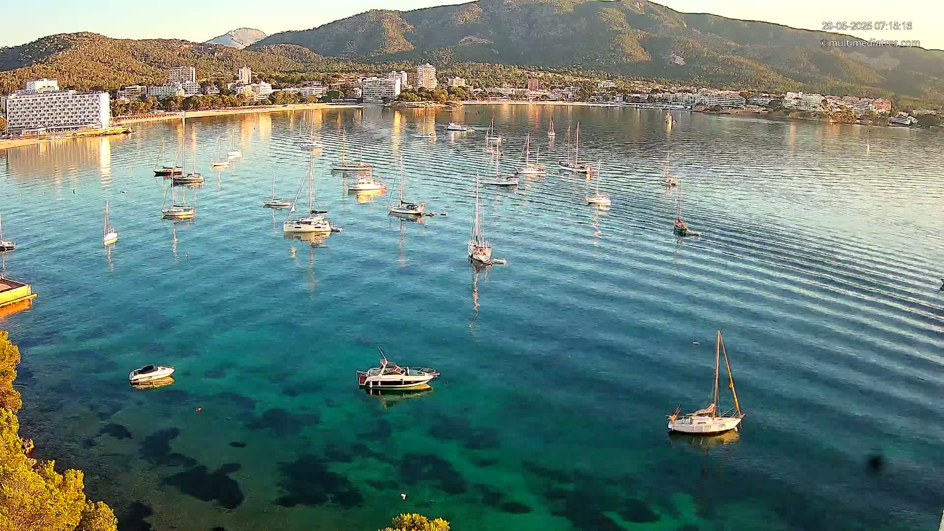 Numerous sailboats and motorboats are anchored in a calm, clear turquoise bay, with a coastal town and mountains visible in the background under a sunny sky.