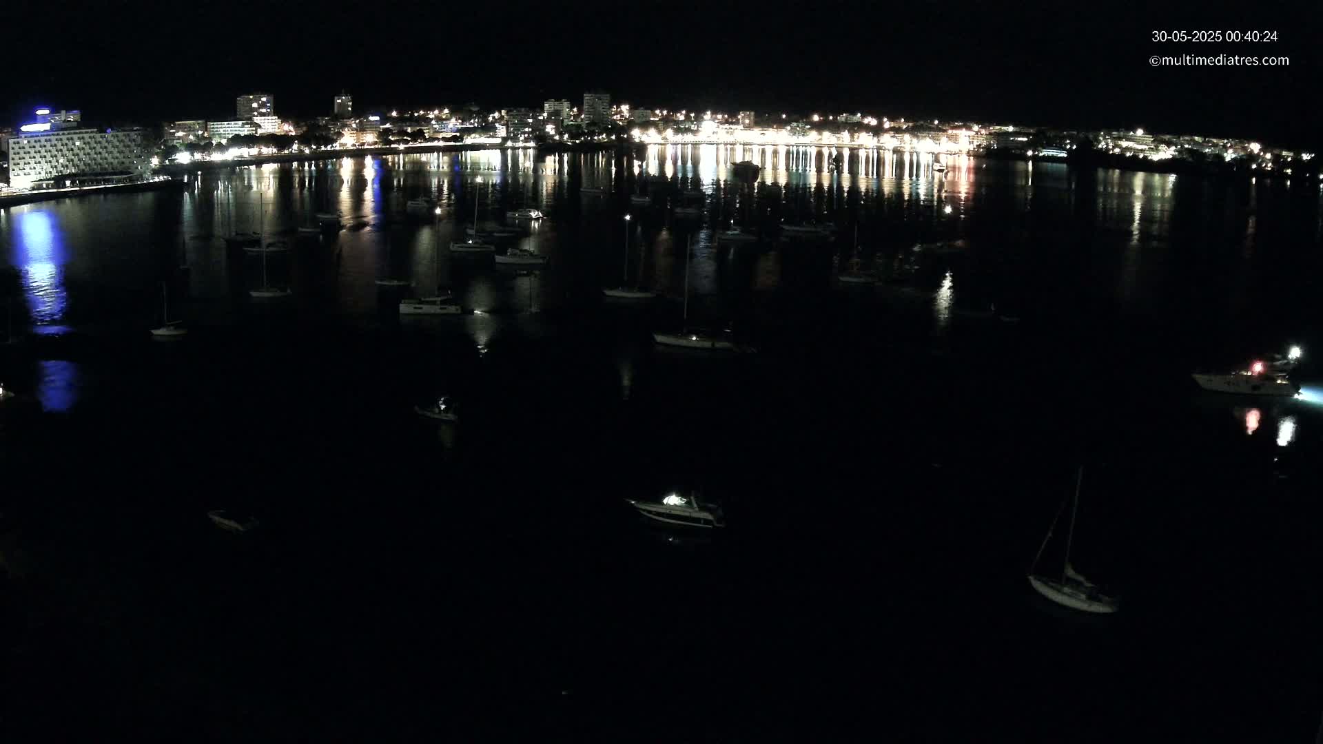At night, numerous boats are moored in a calm body of water in front of a brightly lit city skyline.