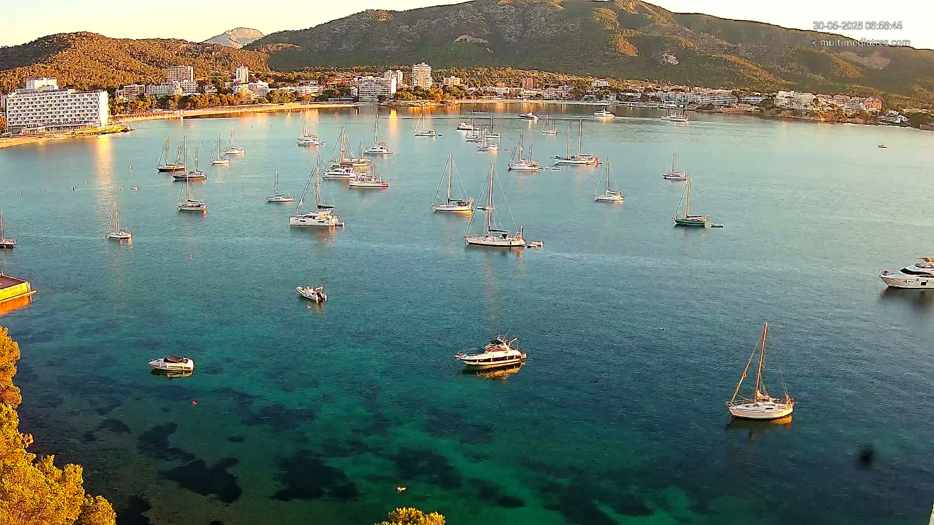 Numerous sailboats and motorboats are anchored in a calm, clear bay next to a coastline with hotels and mountains in the background under a sunny sky.