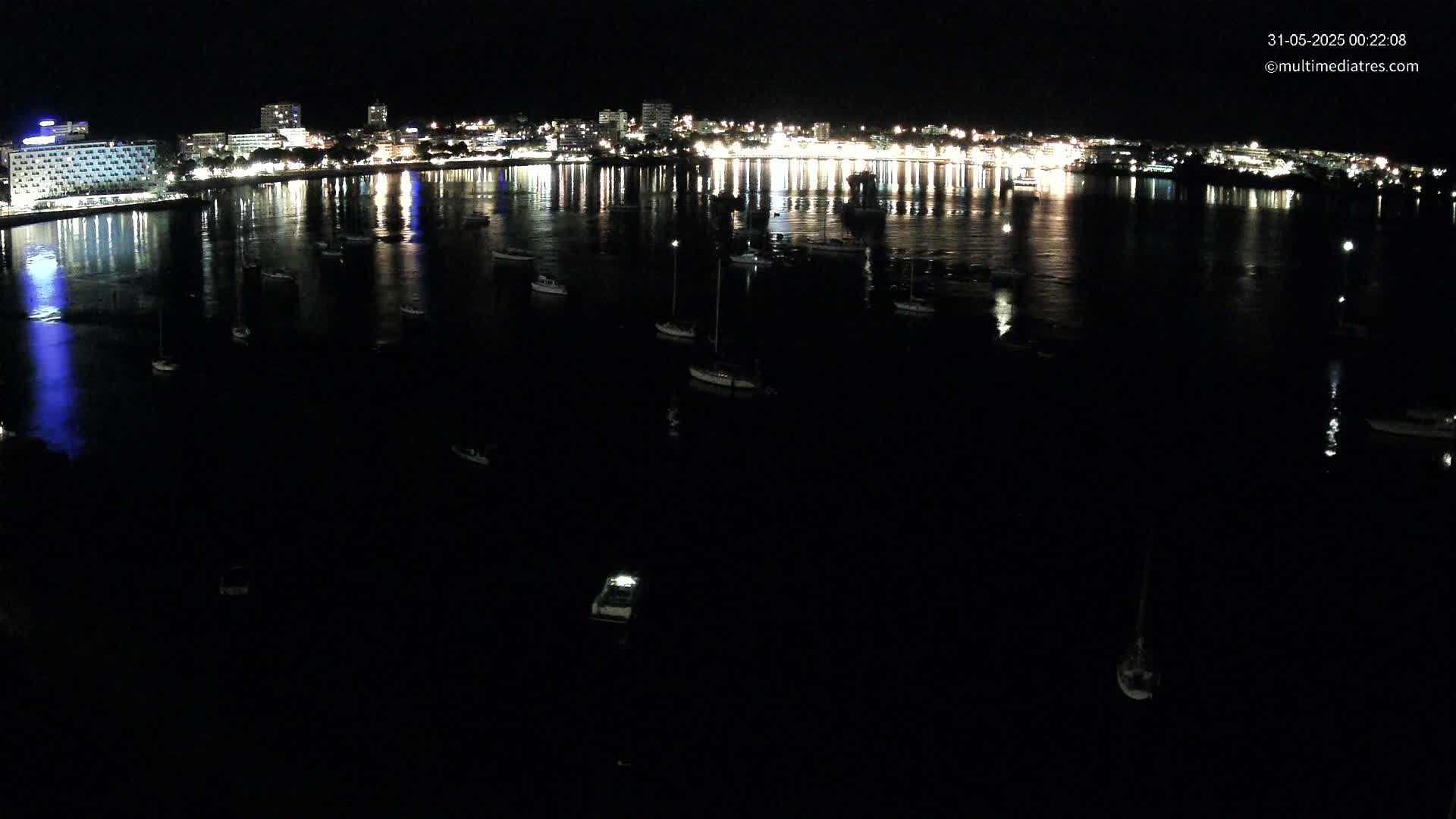 A nighttime view of a city's waterfront, illuminated by lights reflecting on the dark water where several boats are moored.