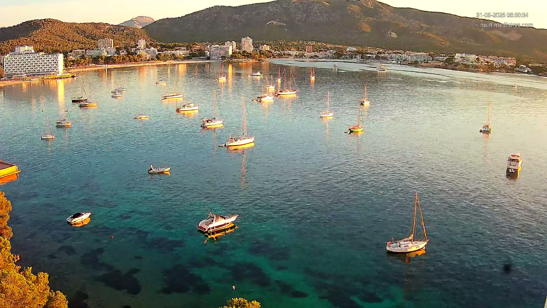 Numerous sailboats and motorboats are moored in a calm, clear bay near a coastal town nestled beneath a mountain range at sunrise.