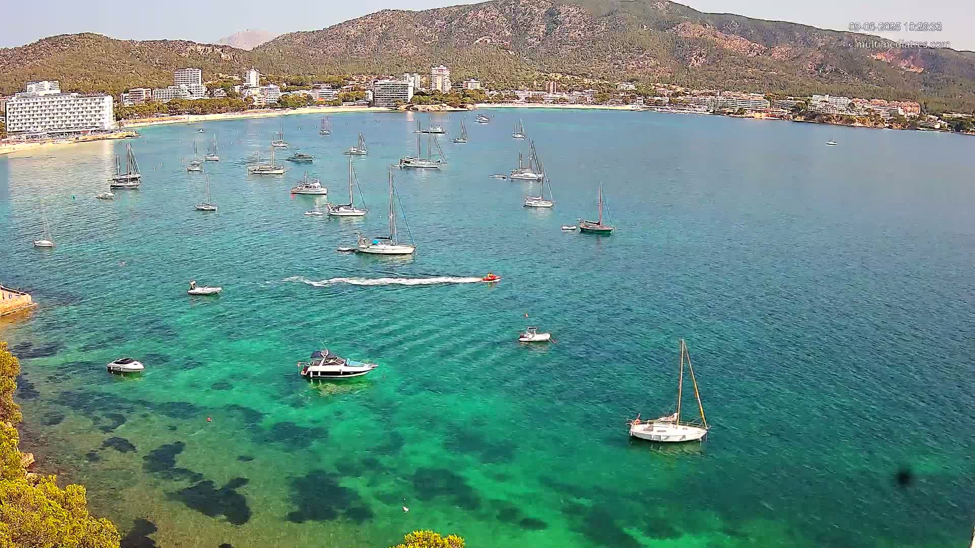 A calm, sunny day shows a bay of clear, turquoise water with numerous sailboats and motorboats anchored near a sandy beach and hotels nestled against a mountain range.