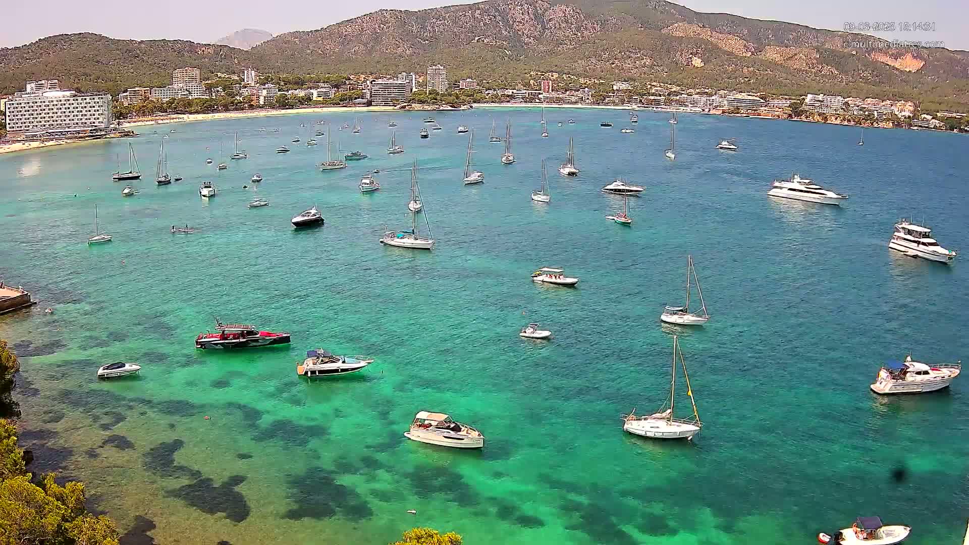 Numerous boats and yachts are anchored in a clear, turquoise bay next to a beach and a mountainous coastline under a sunny sky.