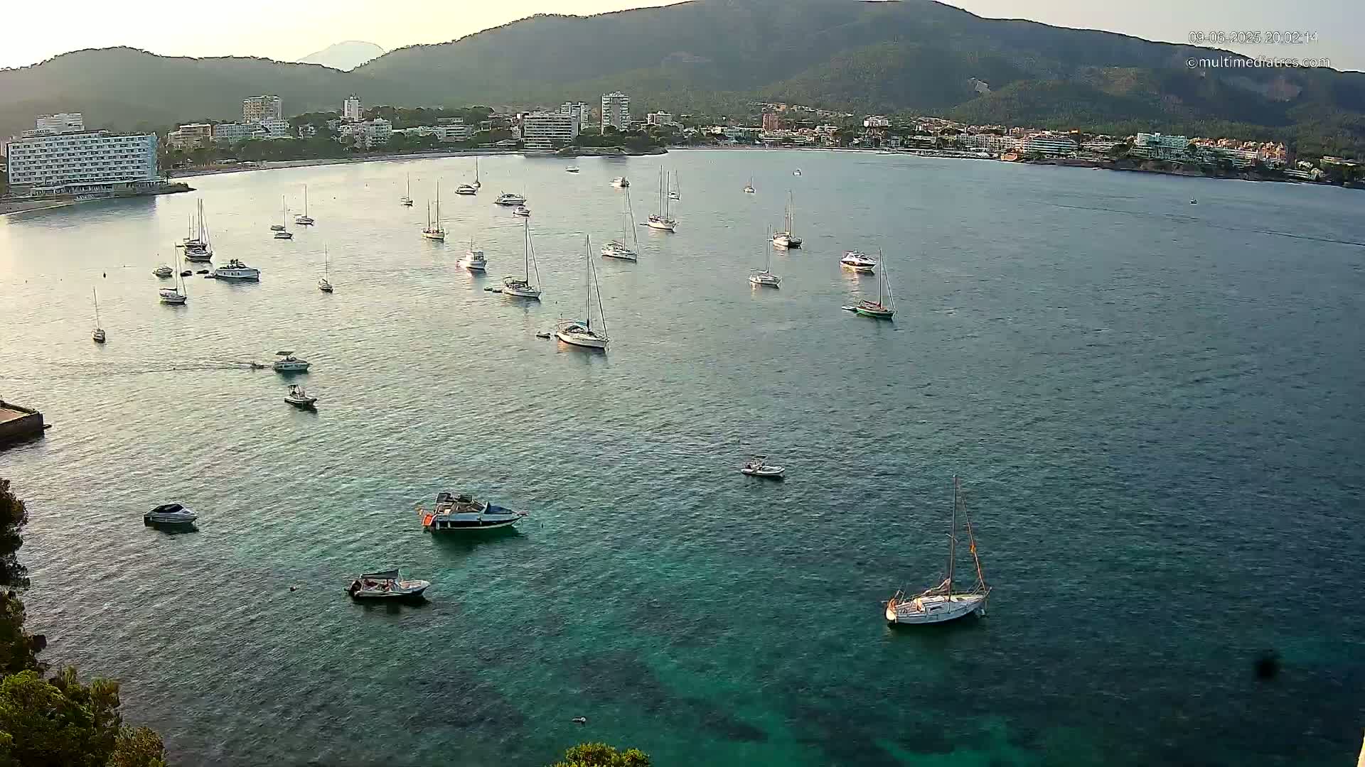 Numerous sailboats and motorboats are scattered across a calm, teal-colored bay, with a mountainous coastline and buildings visible in the background under a clear sky.