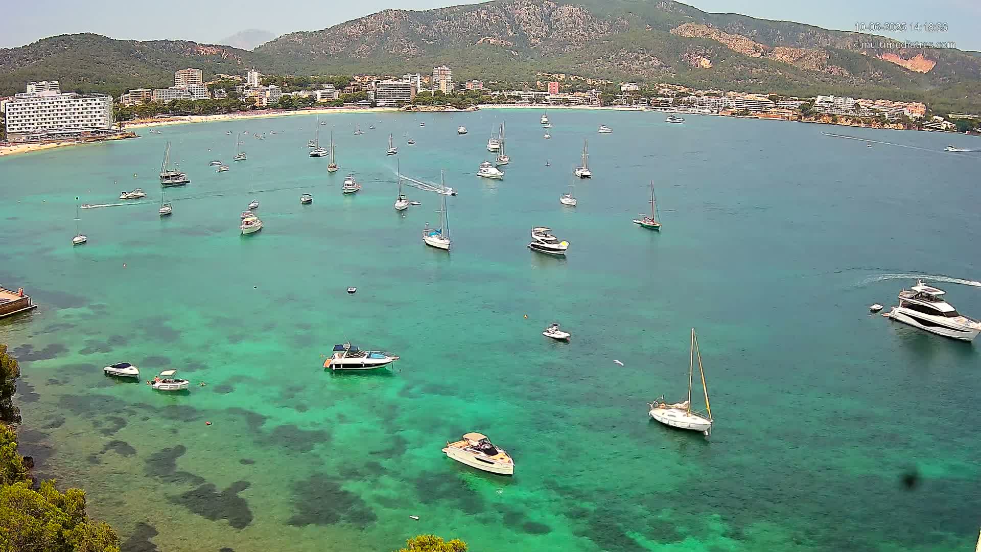 Numerous boats are anchored in a clear, teal bay near a sandy beach and a mountainous coastline under a sunny sky.