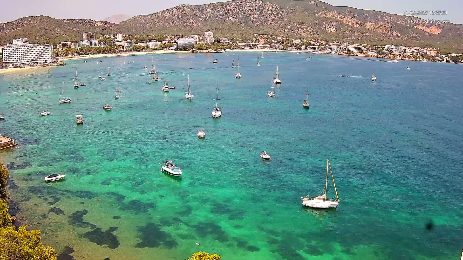 A clear, sunny day shows a calm bay with numerous boats and yachts anchored in vibrant teal water, near a sandy beach and a mountainous backdrop.
