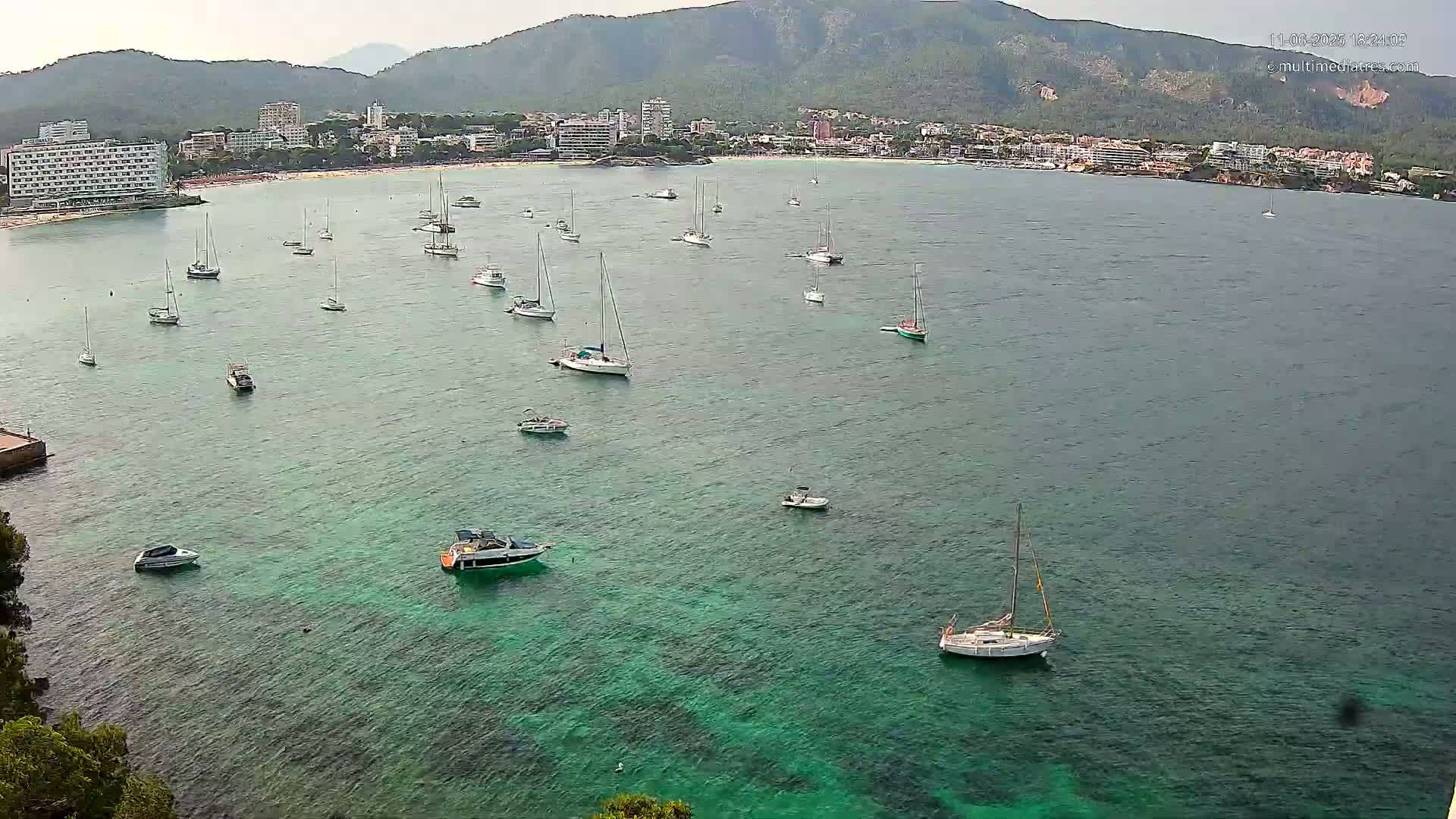 A calm bay with numerous sailboats and motorboats is nestled in front of a coastal town backed by mountains under an overcast sky.
