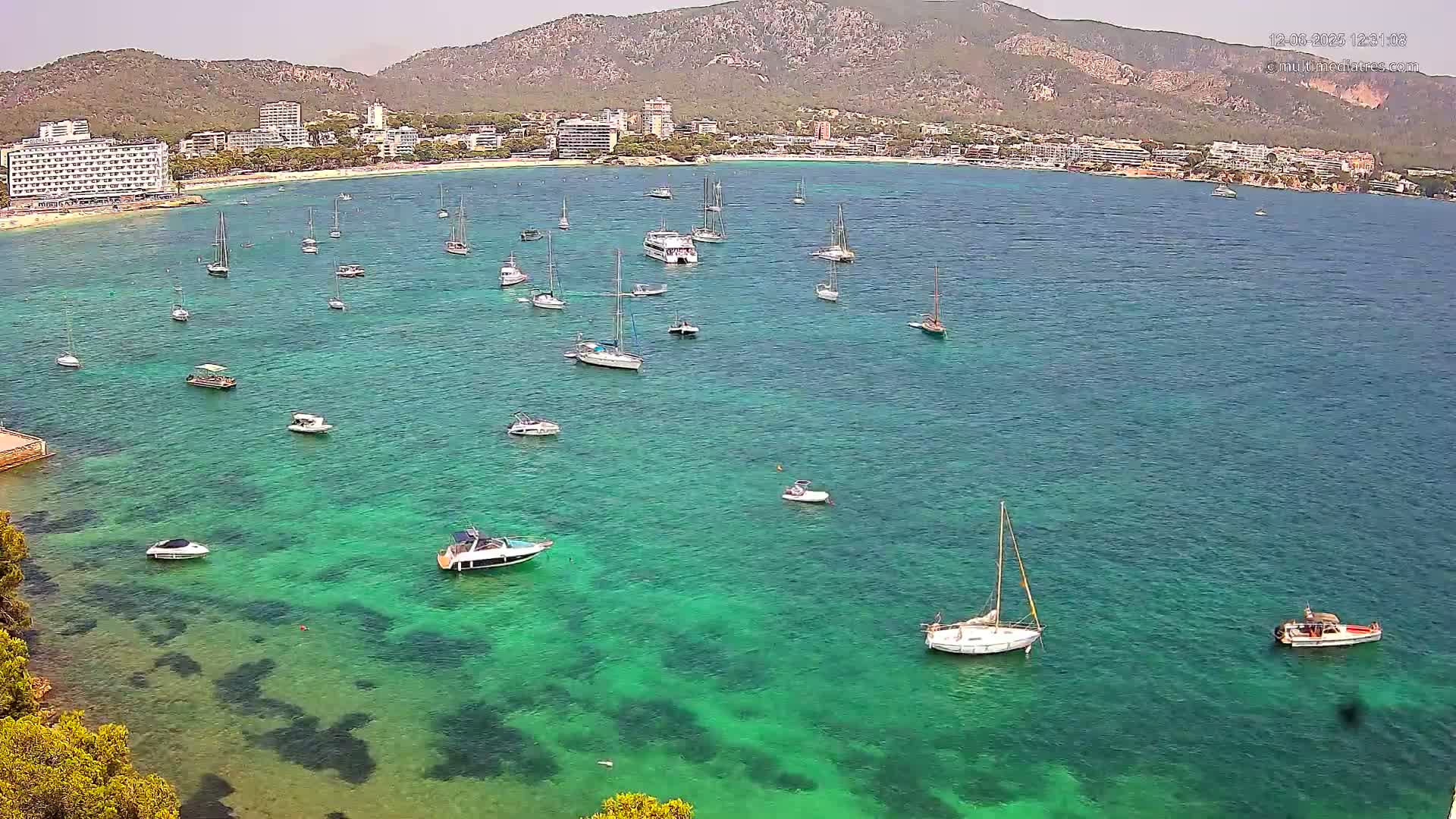 A calm, clear, turquoise bay with numerous boats and sailboats is nestled against a beach and a mountainous backdrop on a sunny day.