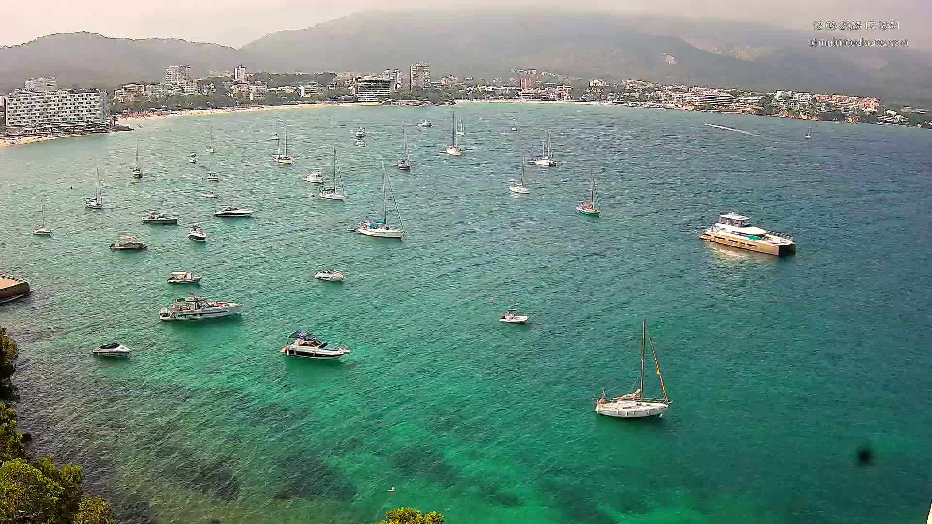 Numerous boats and yachts are anchored in a calm, clear, teal-colored bay near a hazy, mountainous coastline.