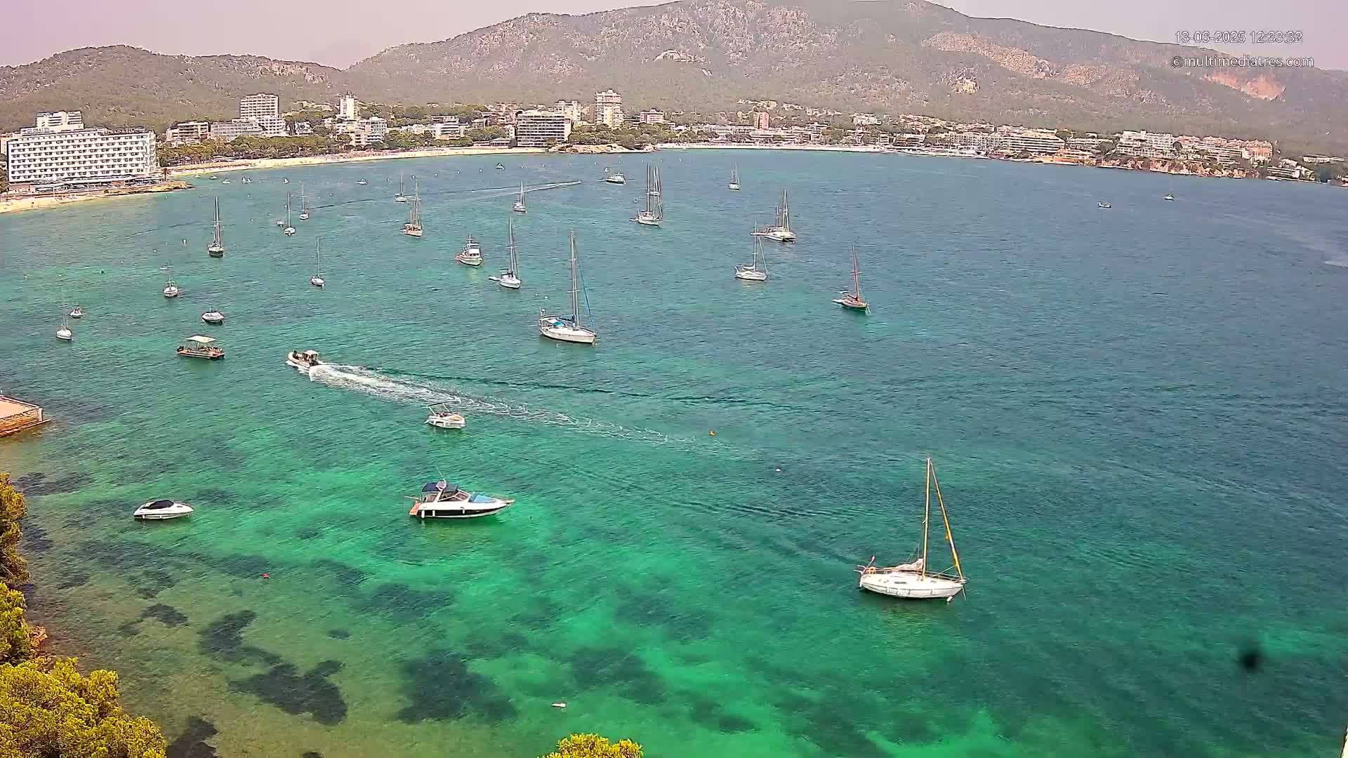 A clear, sunny day reveals numerous boats and sailboats in a calm, bright teal bay, with a shoreline of hotels and buildings backed by a mountain range.