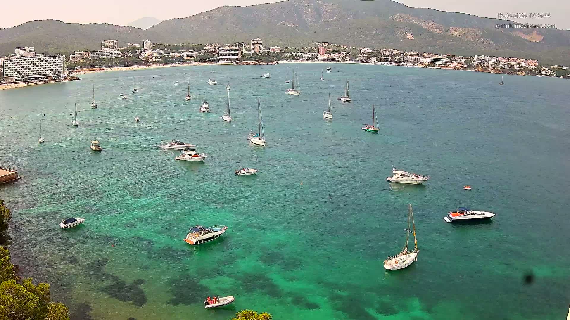 An aerial view shows a calm, clear turquoise bay with numerous sailboats and motorboats anchored near a coastline with hotels and buildings nestled against a mountain range under a hazy sky.