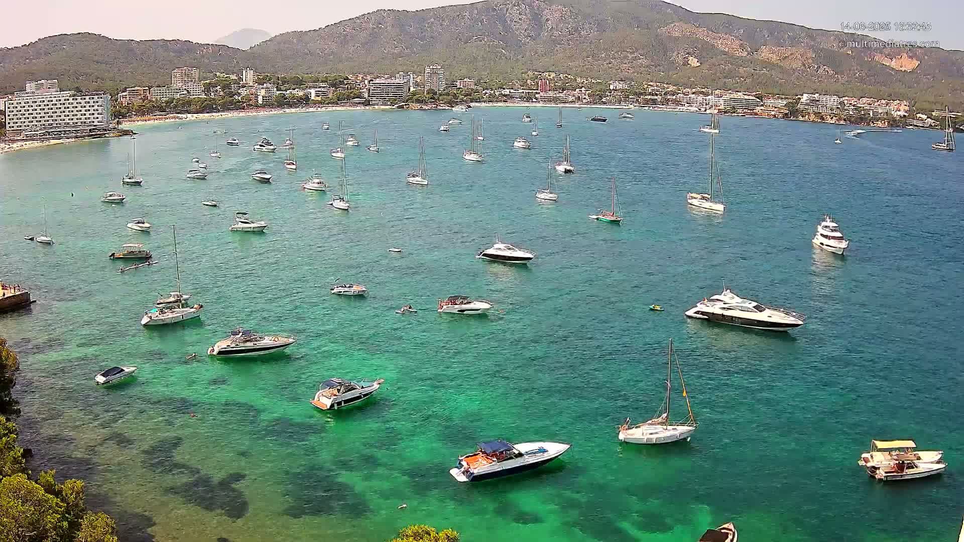 Numerous boats and yachts are anchored in a clear, teal bay on a sunny day, with a mountain range and beach visible in the background.