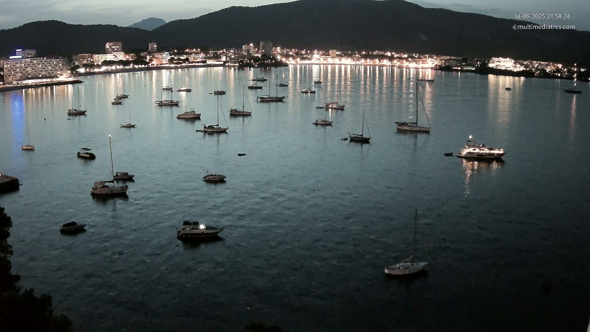 Numerous boats and yachts are anchored in a clear, teal bay on a sunny day, with a mountain range and beach visible in the background.