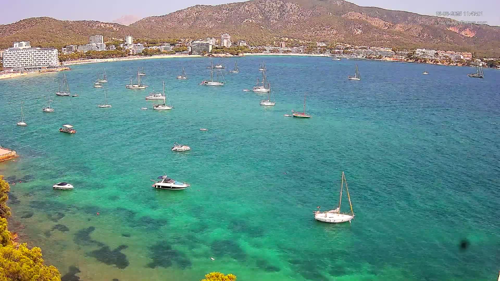 A clear, sunny day shows numerous sailboats and motorboats in a calm, turquoise bay next to a sandy beach and buildings on a hillside.