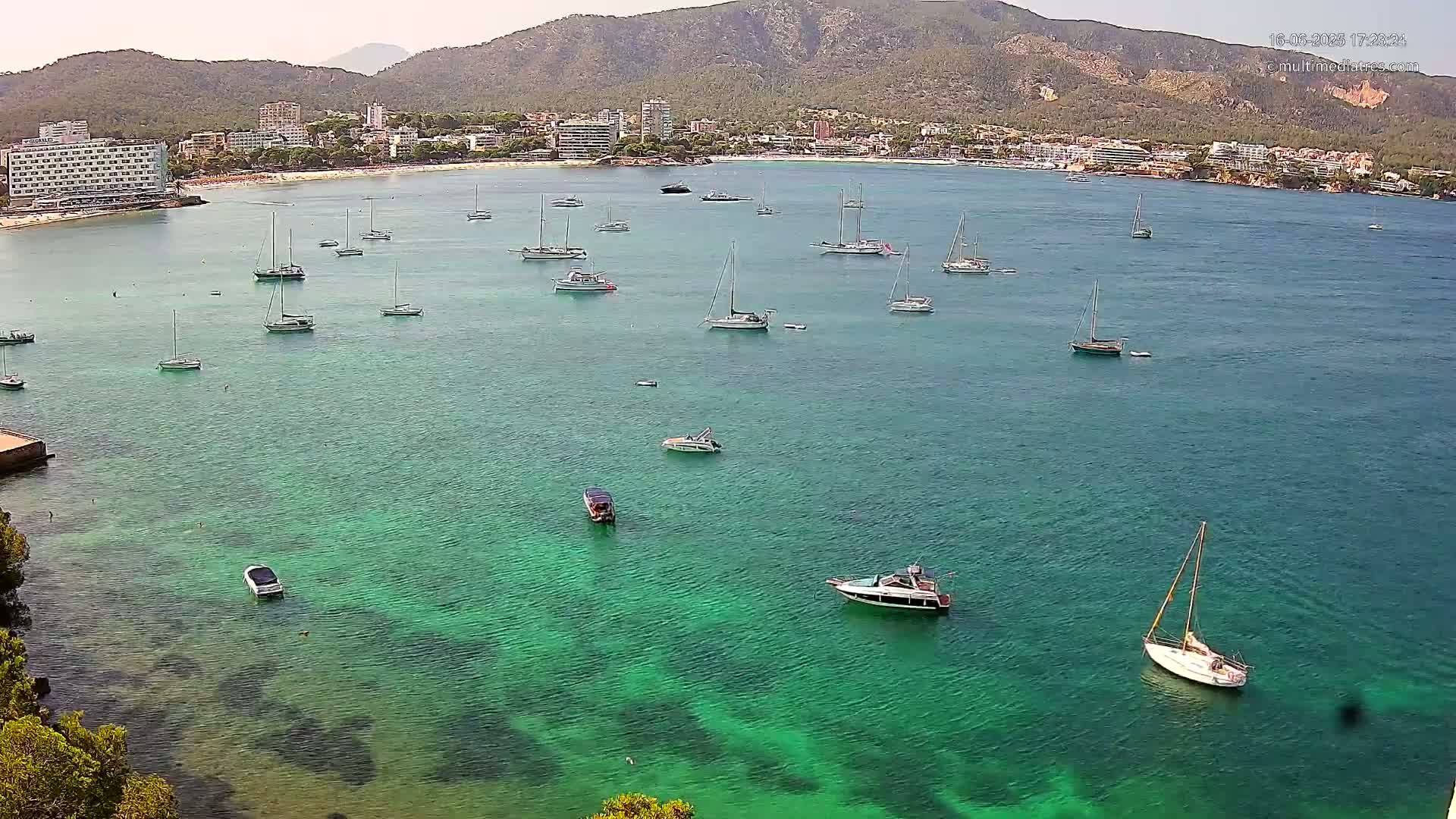 Numerous sailboats and motorboats are anchored in a calm, clear, teal-colored bay, with a sandy beach and hotels visible in the background on a sunny day.