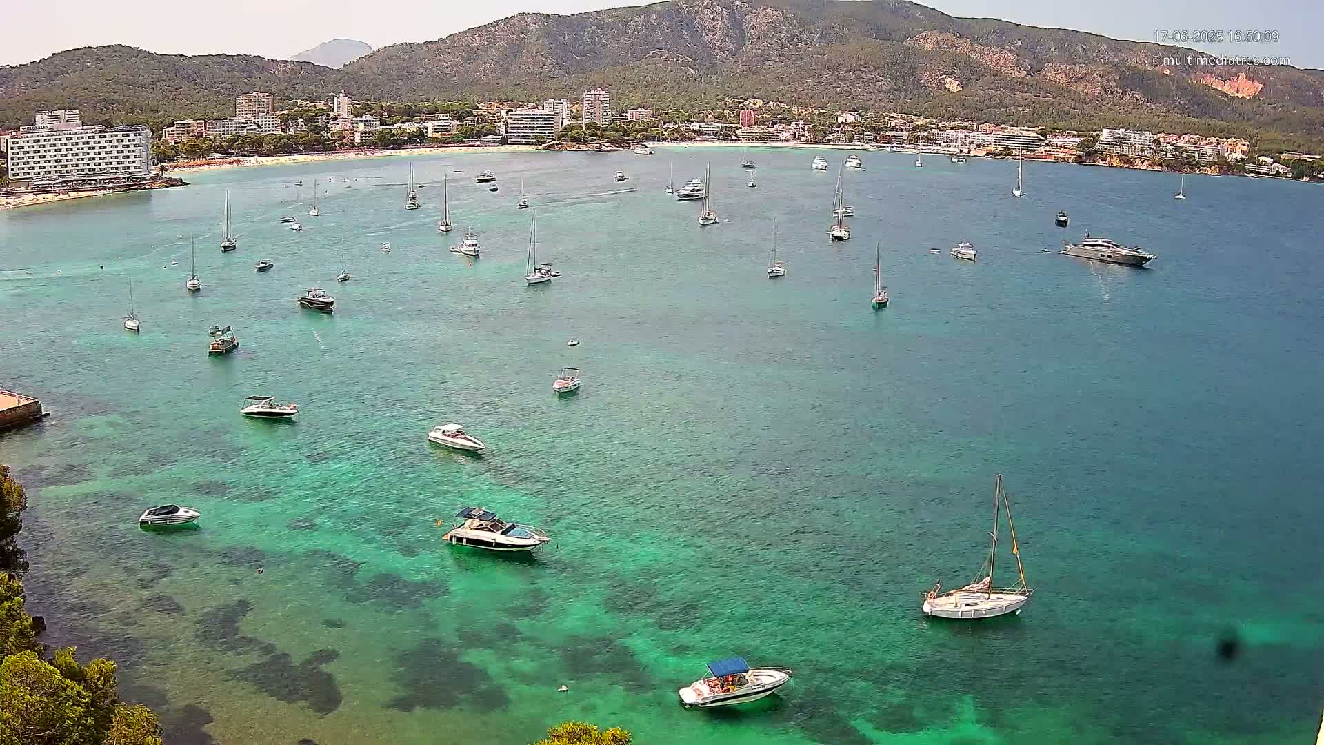 Numerous boats are anchored in a clear, teal bay near a beach and resort town nestled against a mountainous backdrop on a sunny day.