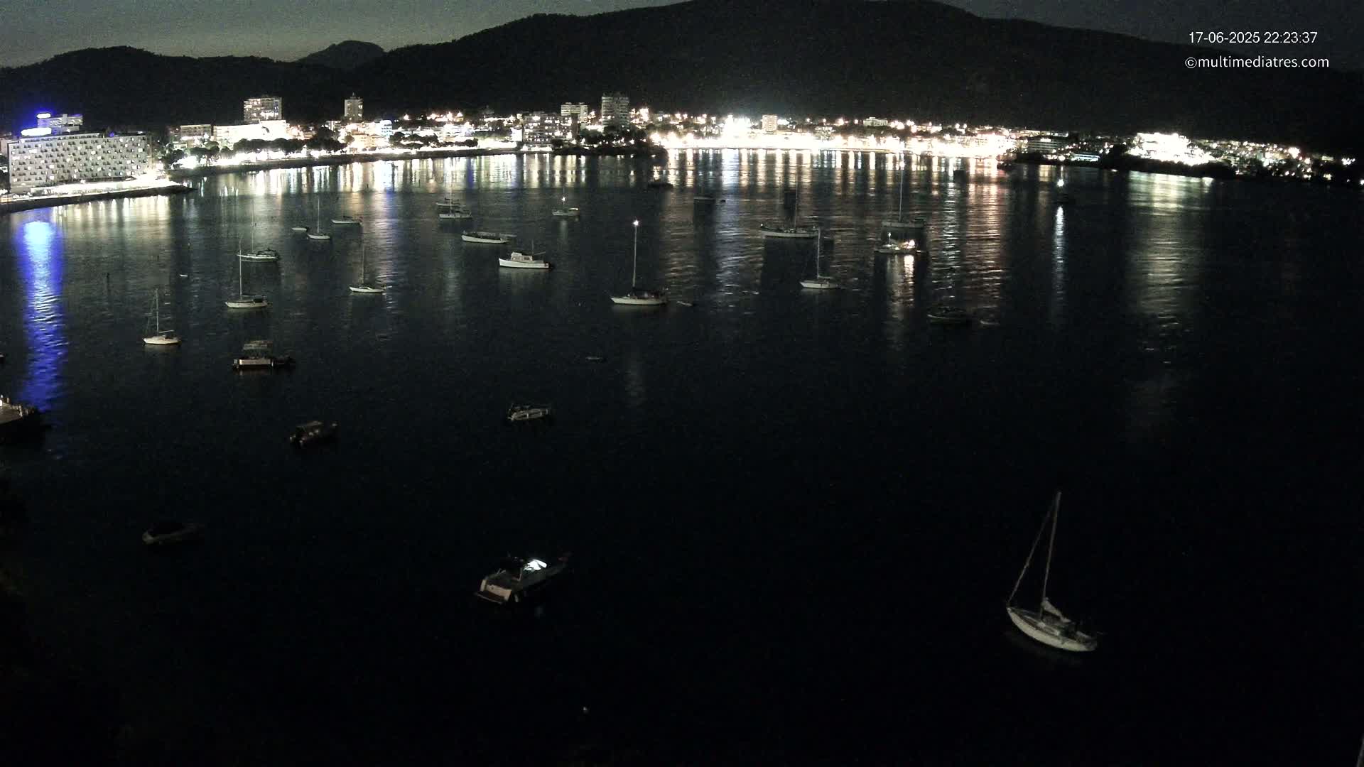 A nighttime view of a calm bay with numerous boats at anchor, reflecting the lights of a city on the shoreline.