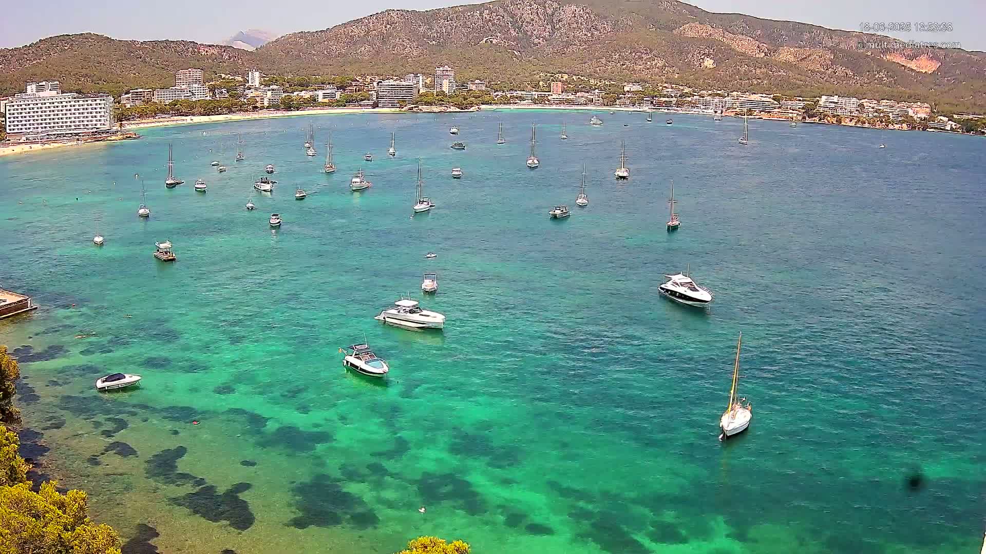 A clear, sunny day reveals a calm, turquoise bay filled with numerous boats, anchored near a sandy beach and hotels nestled against a mountainous backdrop.