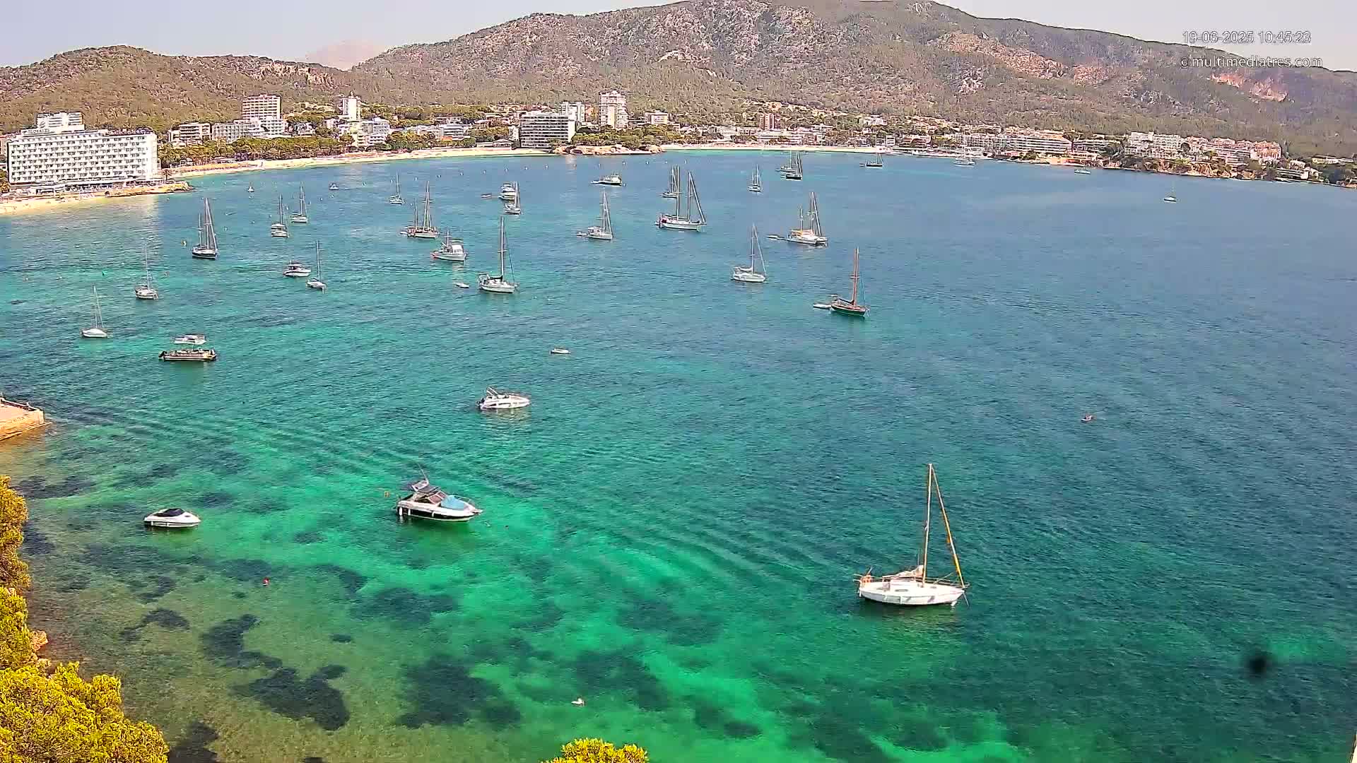 A clear, sunny day shows a calm, bright teal bay with numerous sailboats and motorboats anchored near a sandy beach and shoreline with hotels and buildings nestled against a mountainous backdrop.