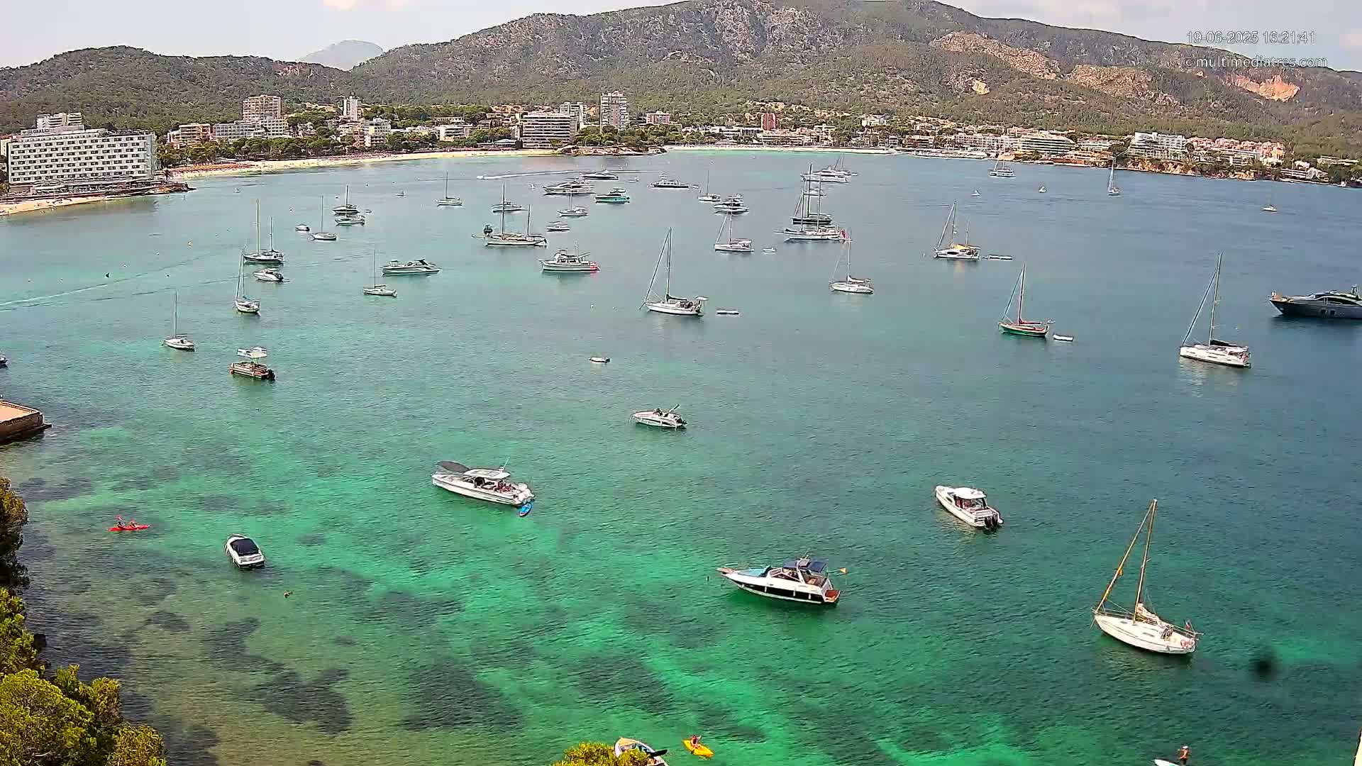 Numerous boats and yachts are moored in a calm, clear, teal-green bay, near a shoreline with buildings and a mountain range in the background under a mostly sunny sky.