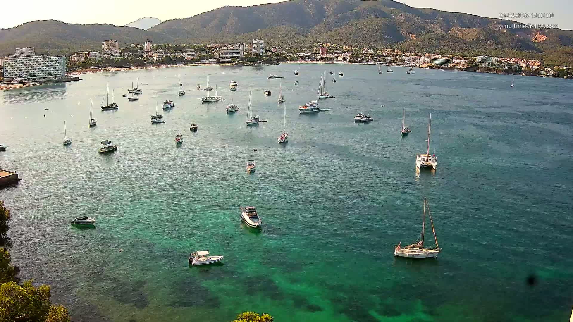 Numerous boats are scattered across a calm, clear, teal bay, nestled against a coastline of buildings and mountains under a bright, sunny sky.