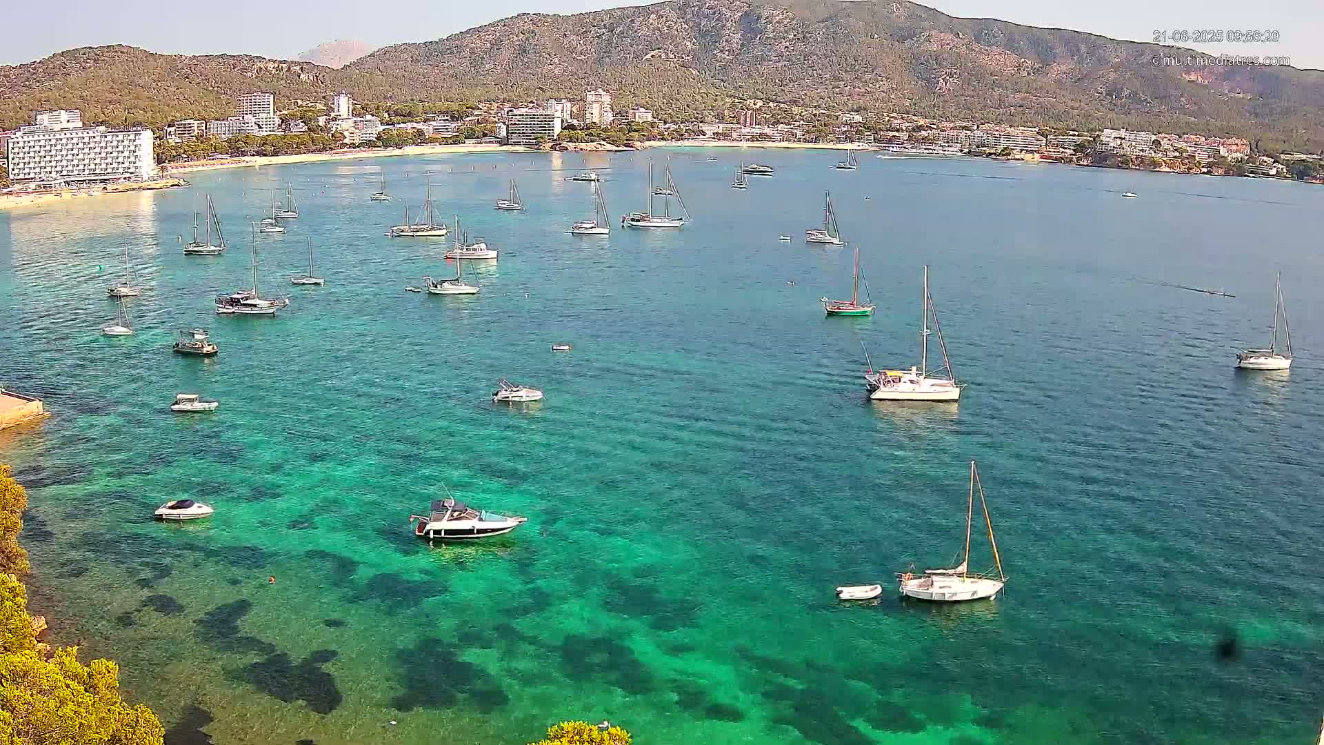 Numerous boats are anchored in a calm, clear, teal-colored bay, next to a sandy beach and hotels on a sunny day, with mountains visible in the background.