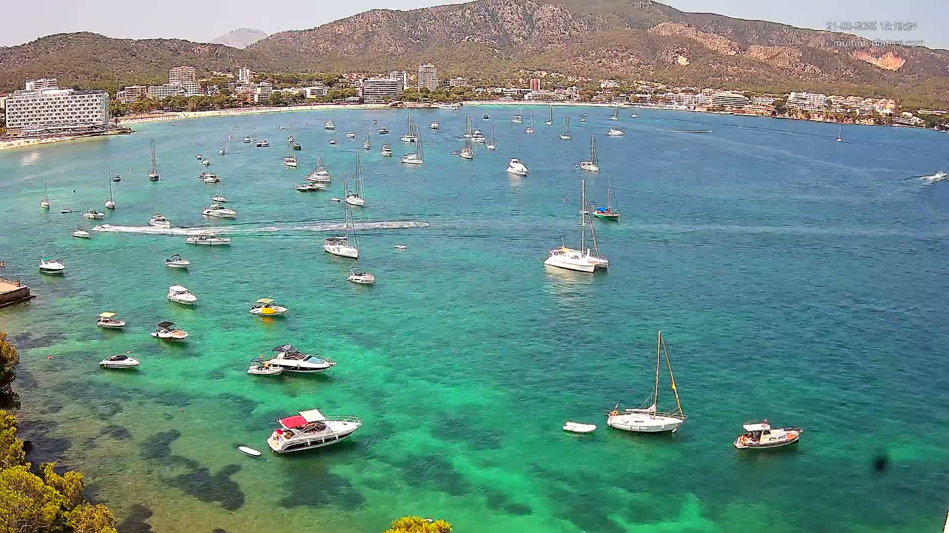 Numerous boats and yachts are anchored in clear, teal water near a beach and mountainous coastline on a sunny day.