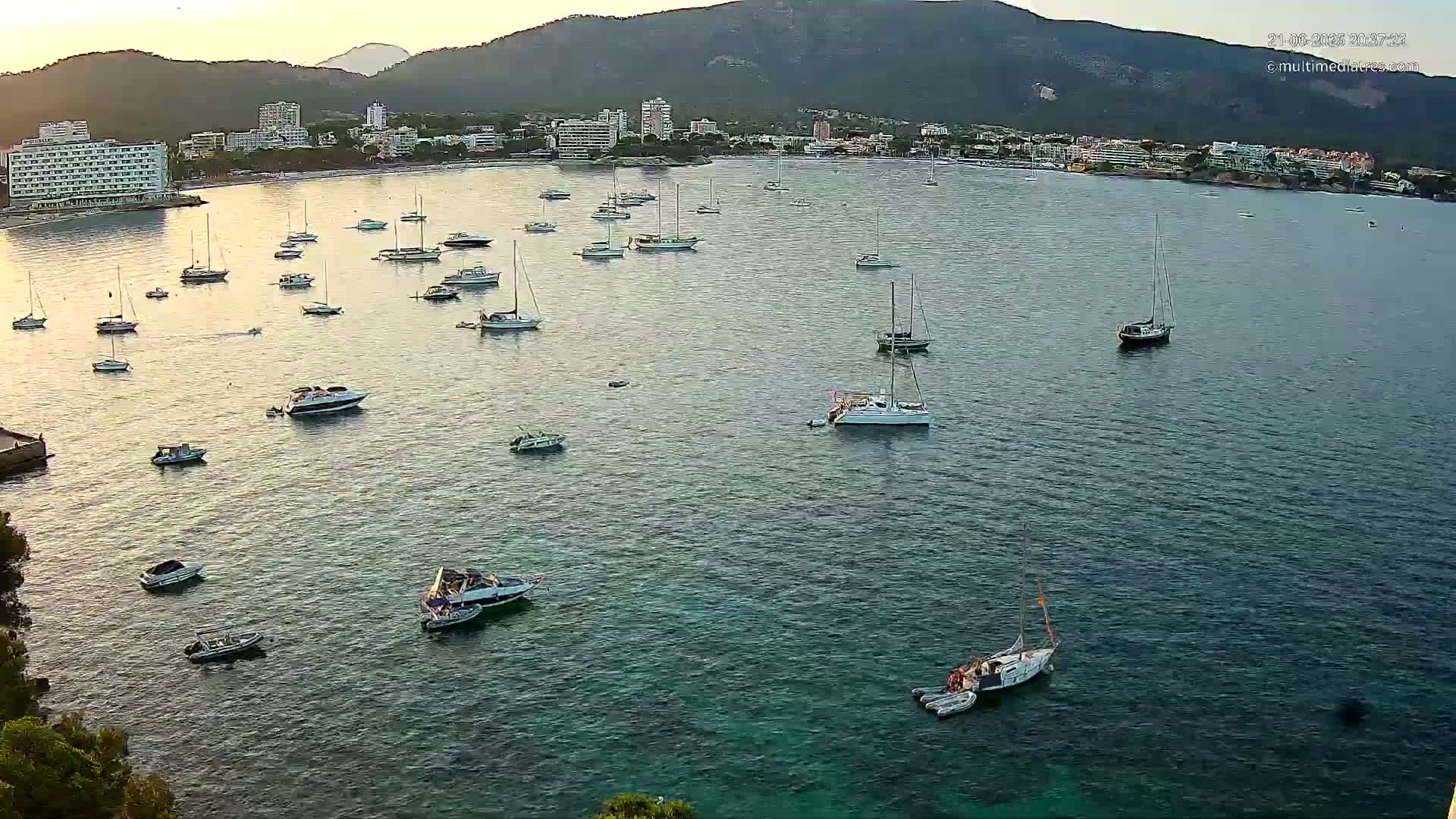 Numerous boats and yachts are moored in a calm bay at sunset, with a mountainous backdrop and a town visible on the shore.