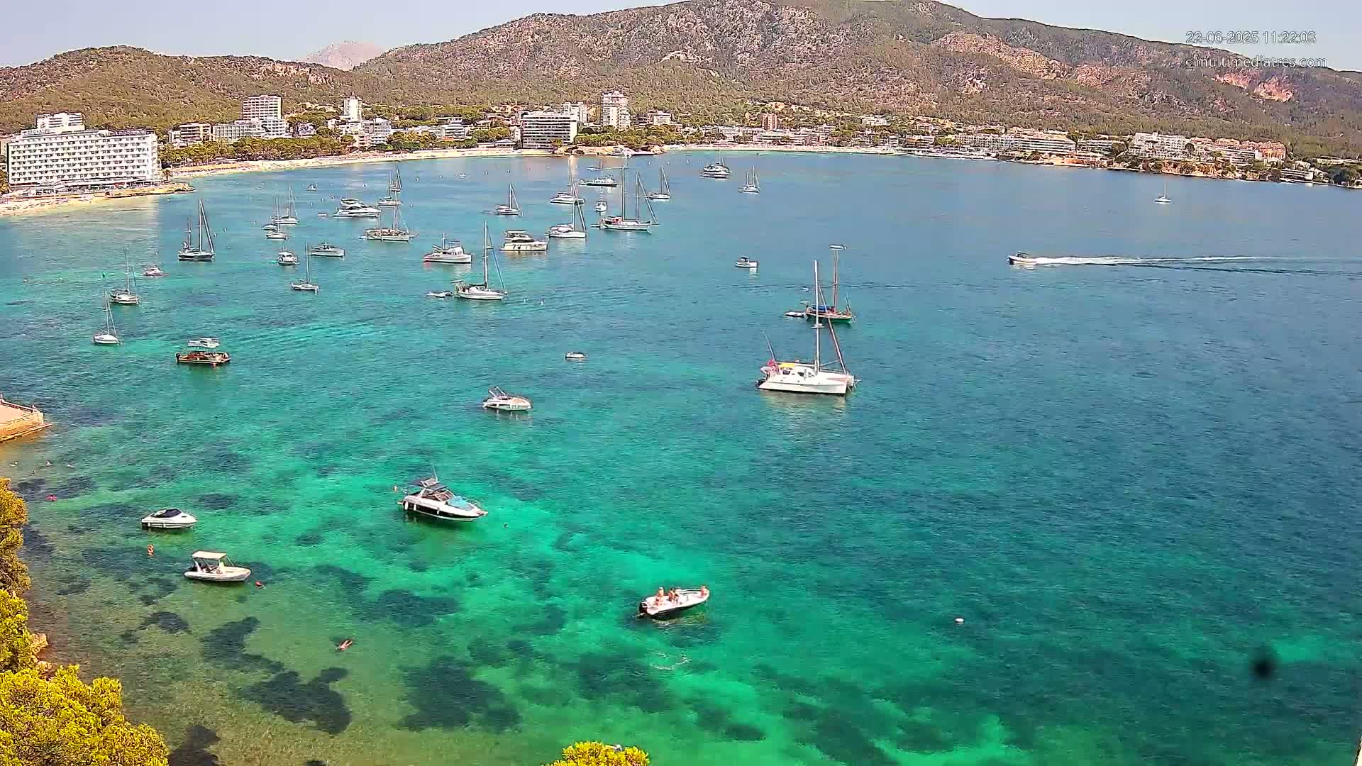 Numerous boats are anchored in a clear, turquoise bay next to a beach and hotels situated at the foot of a mountain range on a sunny day.