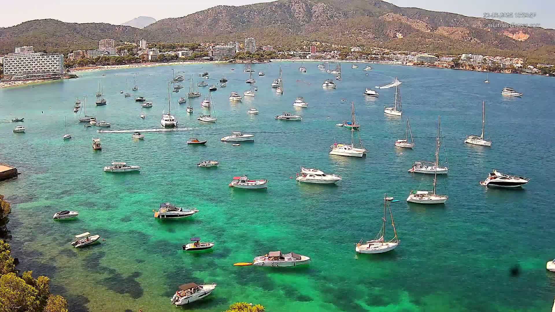 Many boats are anchored in a clear, teal bay next to a mountainous coastline on a sunny day.
