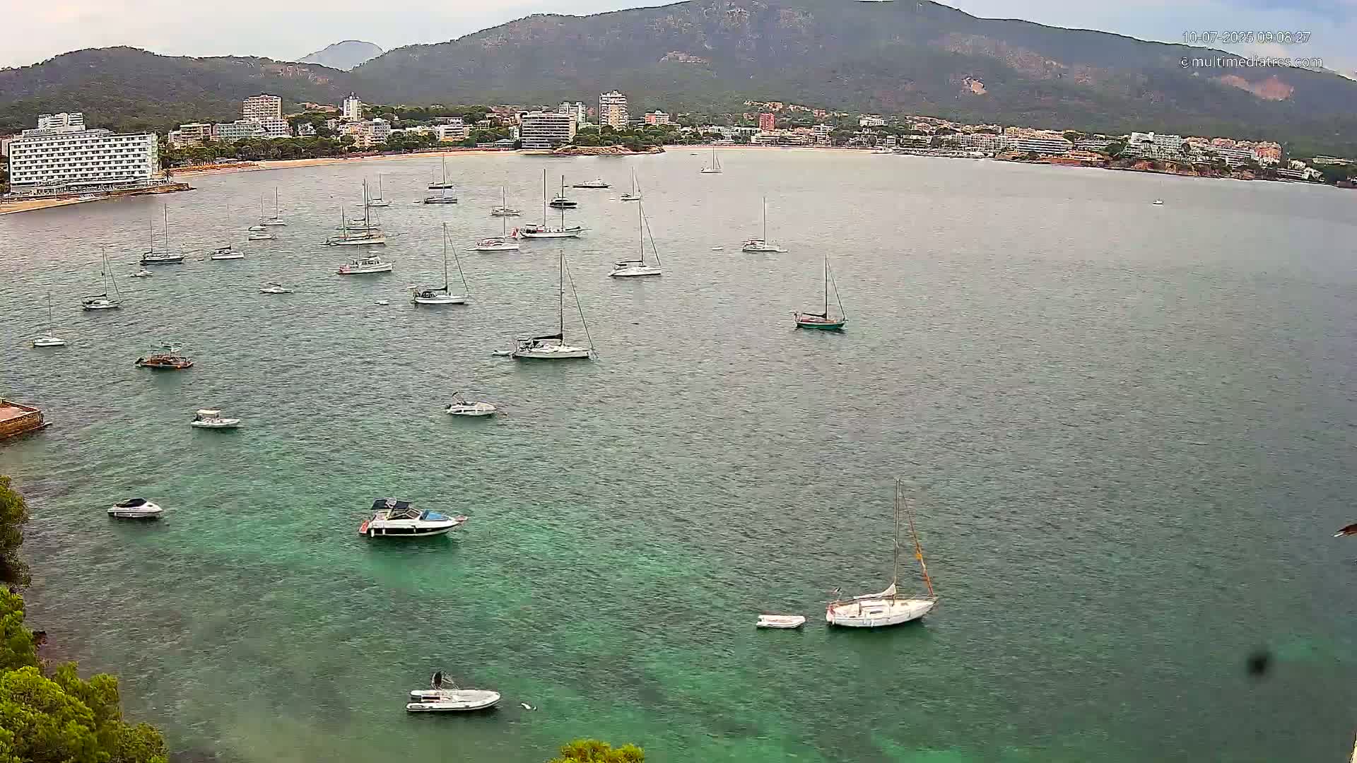 Numerous sailboats and motorboats are anchored in a calm, green bay, with a distant shoreline of buildings and mountains under an overcast sky.
