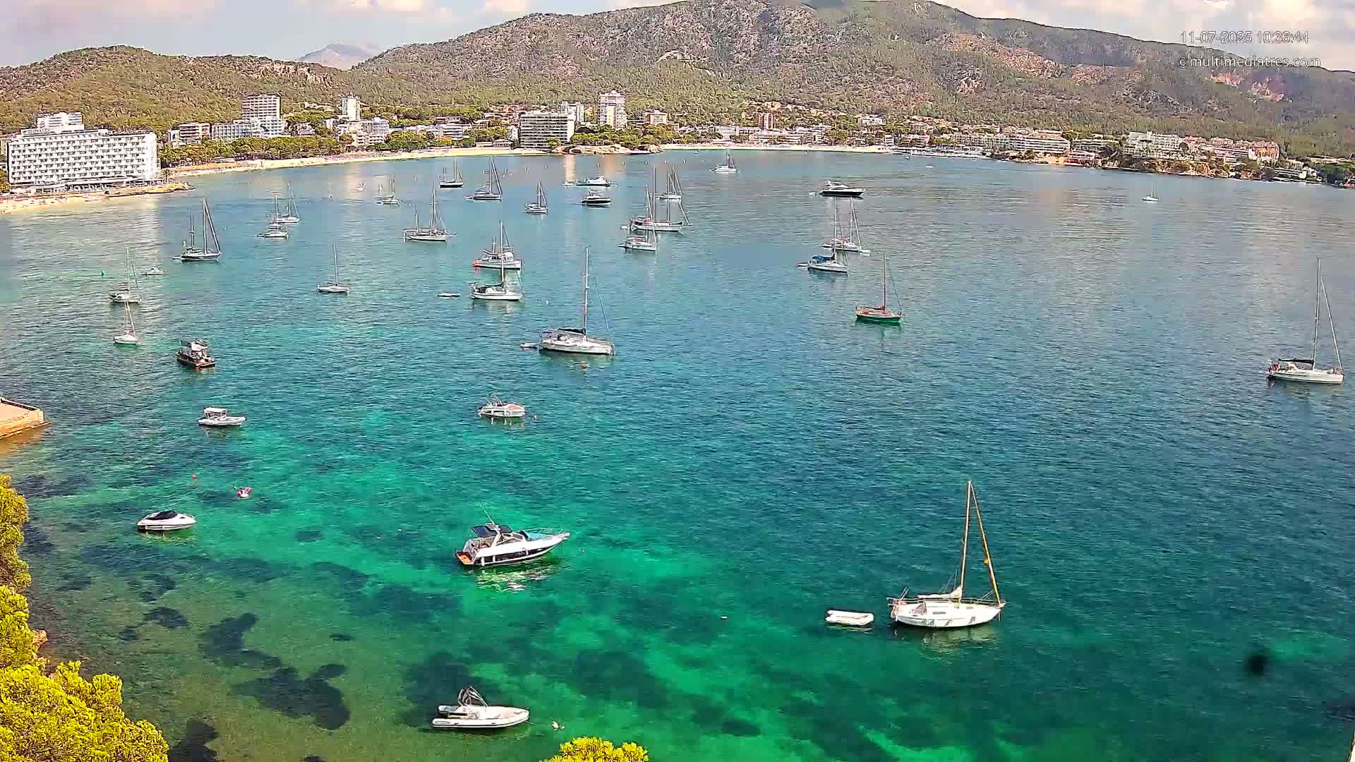 Numerous boats and yachts are anchored in a calm, clear turquoise bay, with a mountain range and hotels visible in the background under a partly cloudy sky.