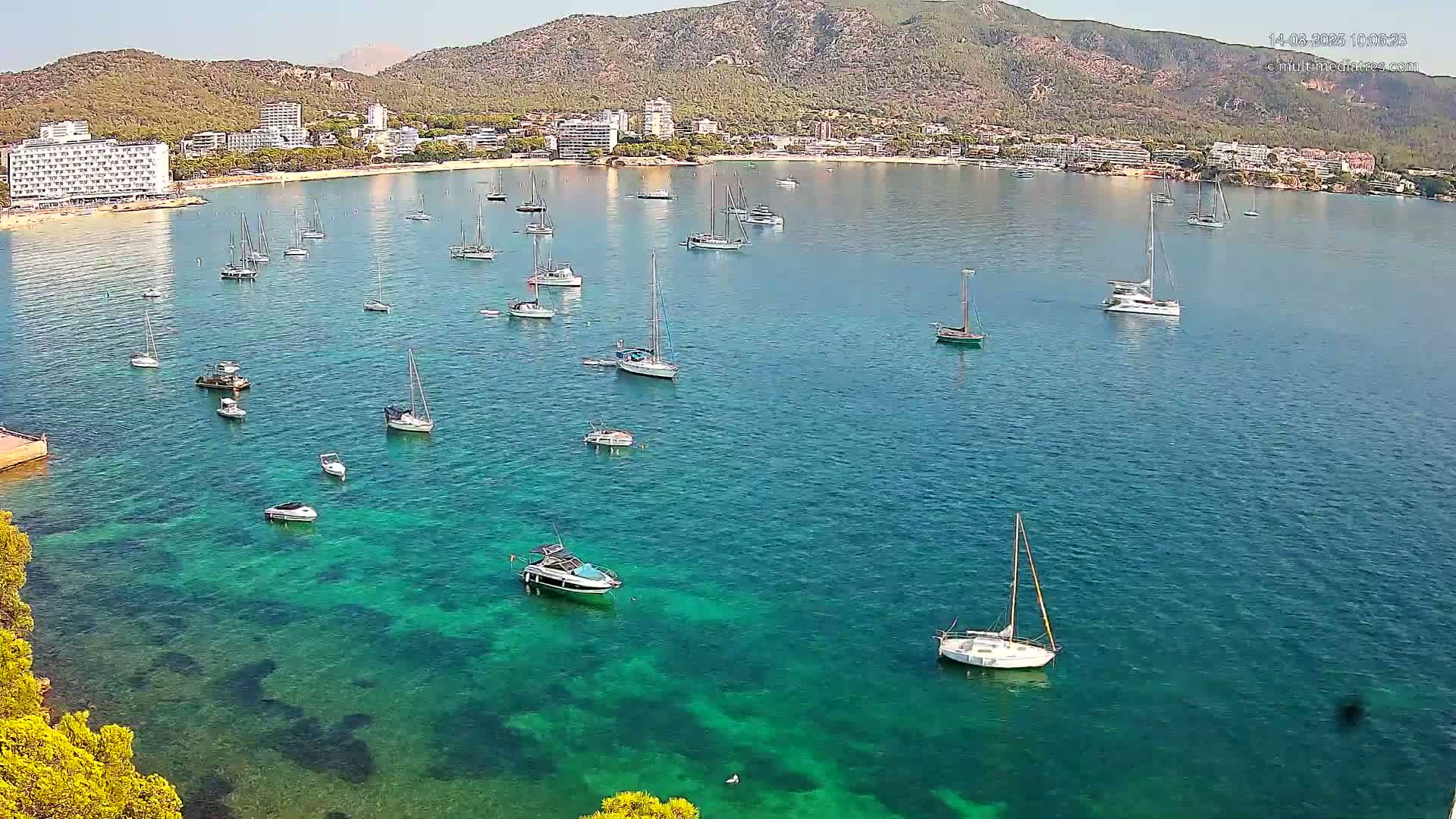 Numerous boats are anchored in a calm, clear, turquoise bay, with a distant shoreline featuring hotels and a mountain under a sunny sky.