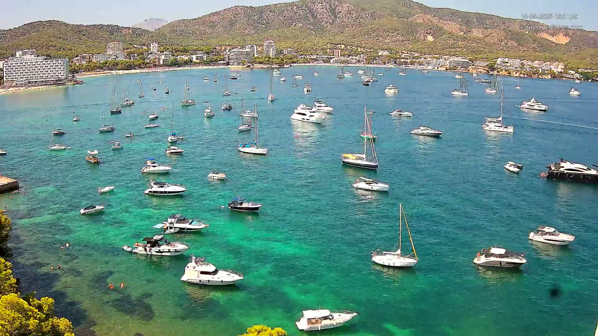 Numerous boats are anchored in a calm, clear, turquoise bay, with a distant shoreline featuring hotels and a mountain under a sunny sky.