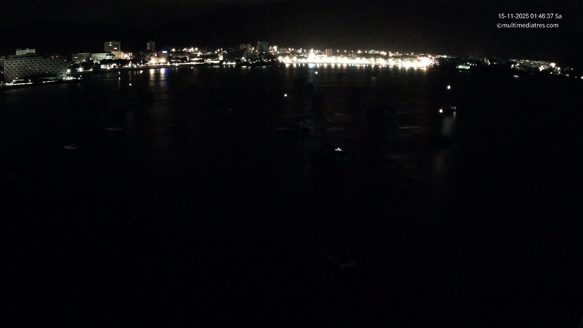 A nighttime view reveals a distant city skyline illuminated by numerous lights reflecting on a calm body of water under a clear sky.
