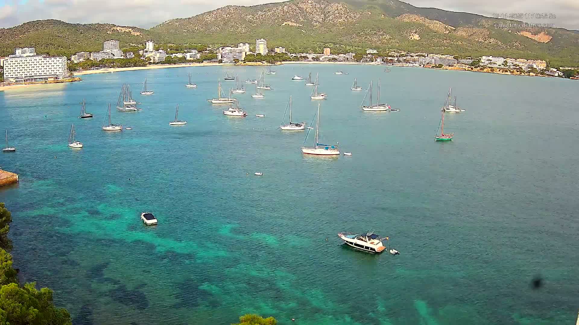 A wide coastal bay with remarkably clear, turquoise water is filled with many sailboats and some motorboats, bordered by a resort town with a sandy beach and lush mountains under a partly cloudy sky.