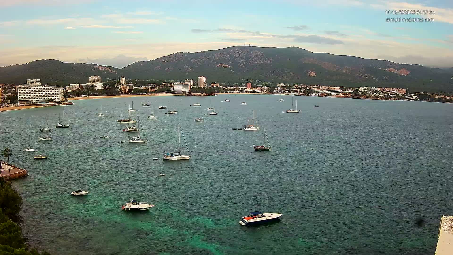 Numerous sailboats and a few motorboats dot a sunny, turquoise bay fronting a coastal town with a sandy beach and lush mountains, all under a blue sky dotted with fluffy white clouds.