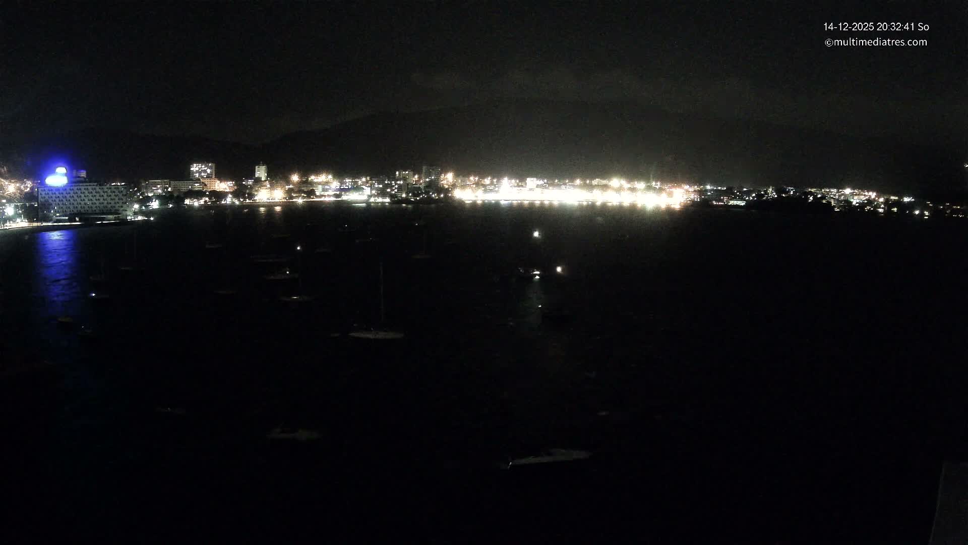 Numerous sailboats and a few motorboats dot a sunny, turquoise bay fronting a coastal town with a sandy beach and lush mountains, all under a blue sky dotted with fluffy white clouds.
