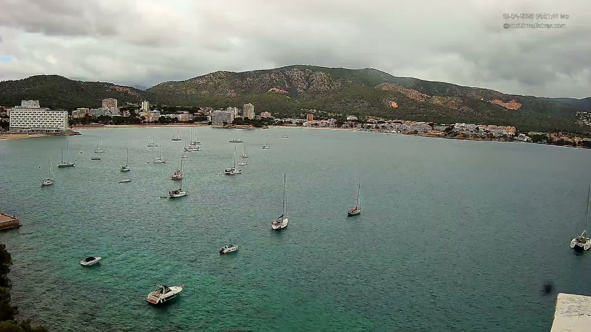 Numerous sailboats and a few motorboats dot a sunny, turquoise bay fronting a coastal town with a sandy beach and lush mountains, all under a blue sky dotted with fluffy white clouds.